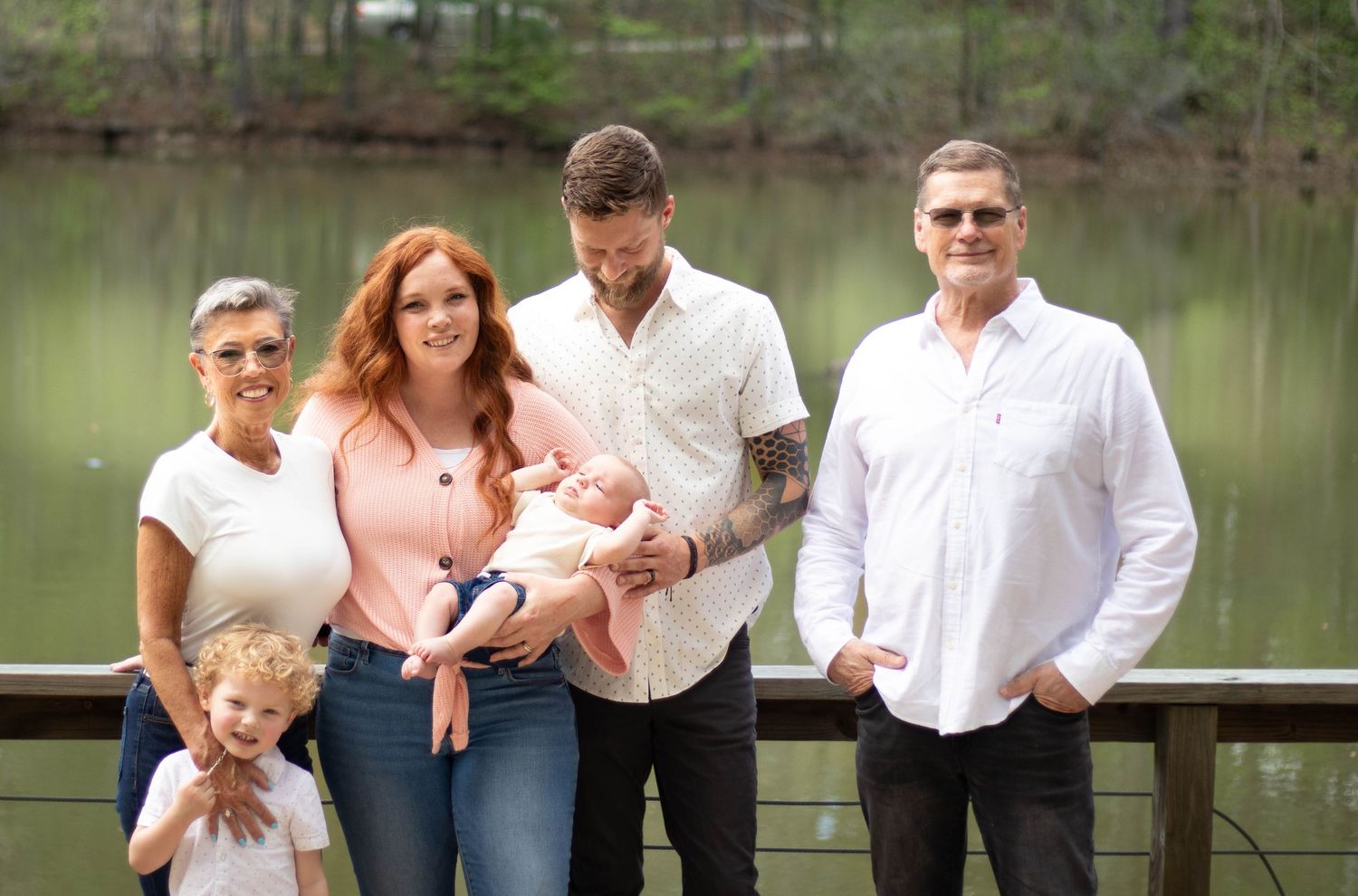 A multigenerational family poses together near a pond, including parents holding a newborn baby.