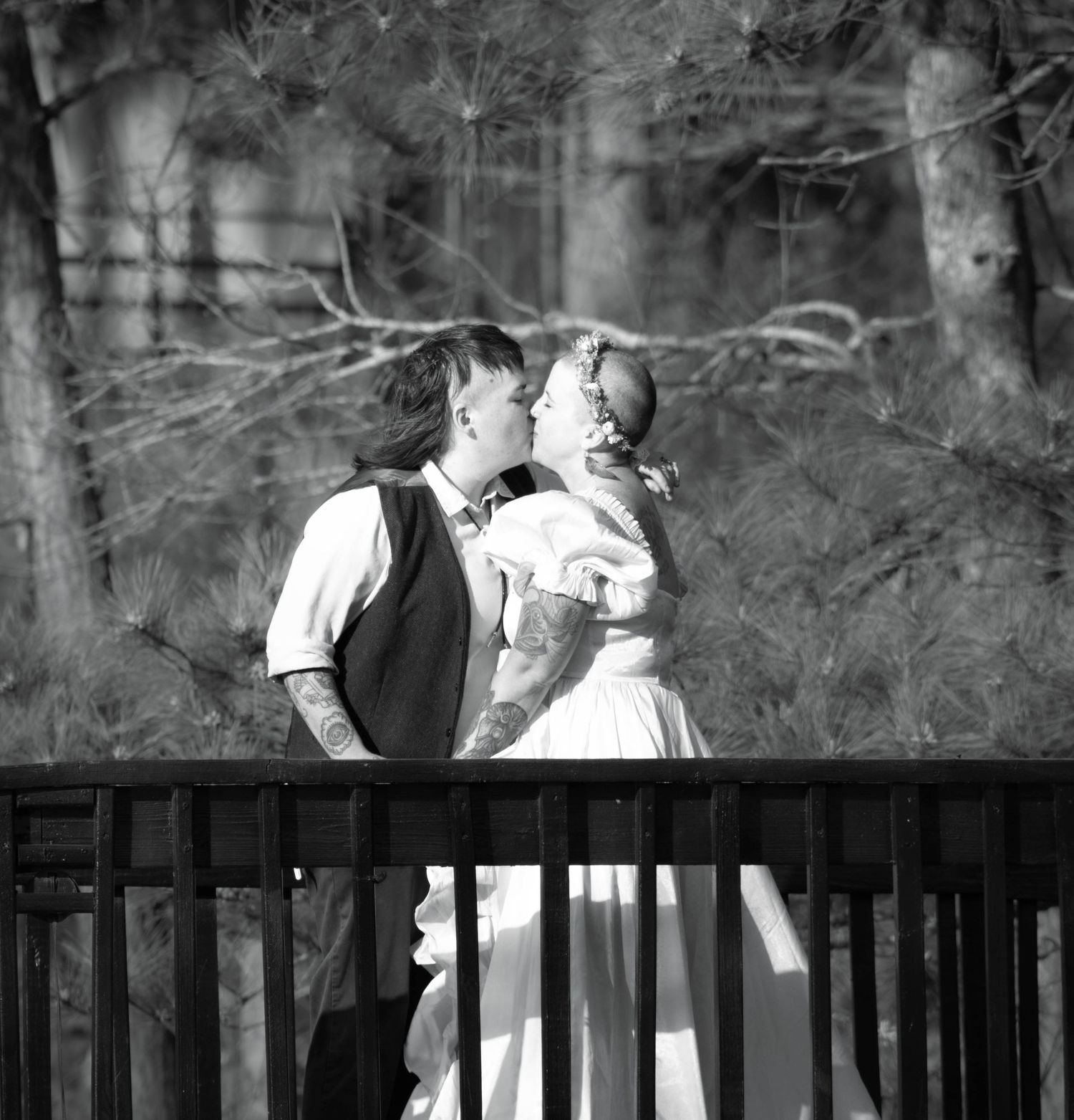 Black and white photo of a couple sharing a kiss in wedding attire on a wooden bridge surrounded by trees.