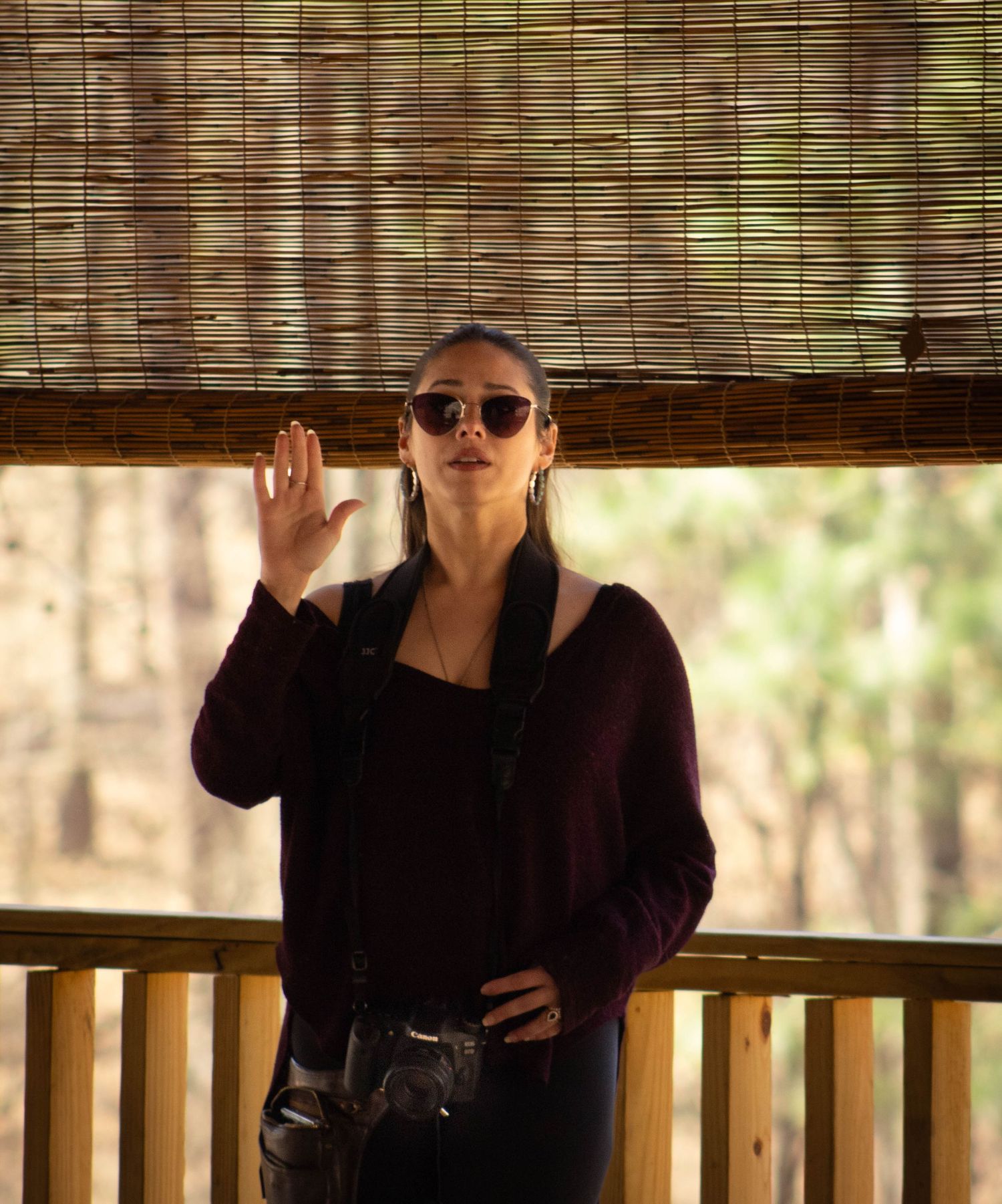 A stylish woman in sunglasses raises her hand while standing on a wooden porch outdoors.