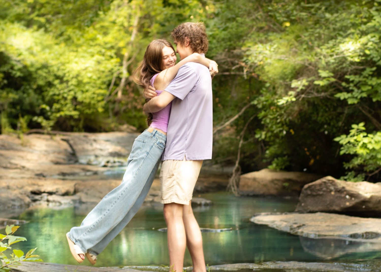 Young couple embracing and hugging romantically by a serene creek surrounded by lush green trees outdoors.