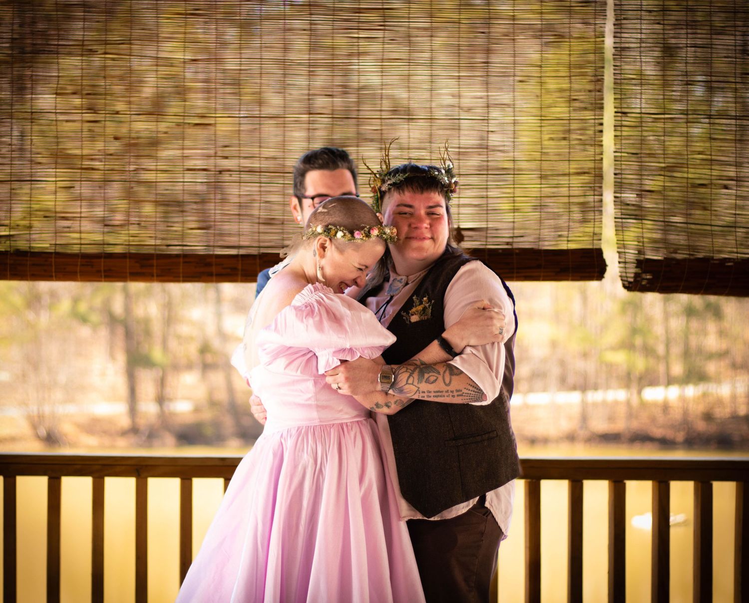 Couple in costume on a porch, man in vest hugging woman in pink dress, with bamboo blinds backdrop.