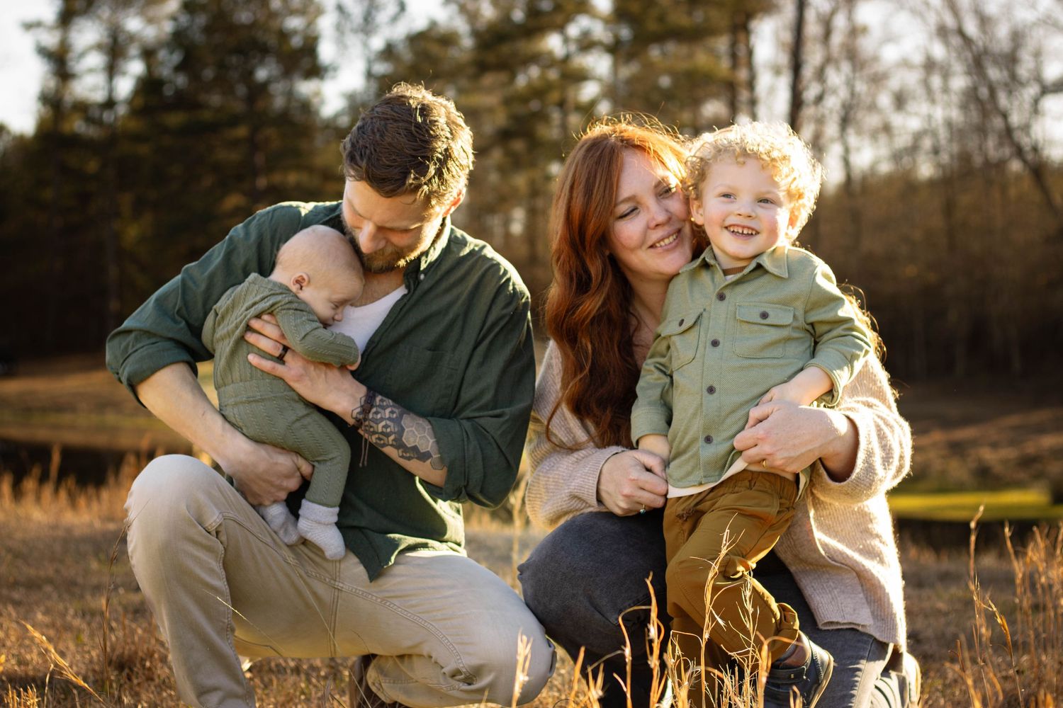Happy family of four with newborn and toddler smiling together in golden sunlit field.