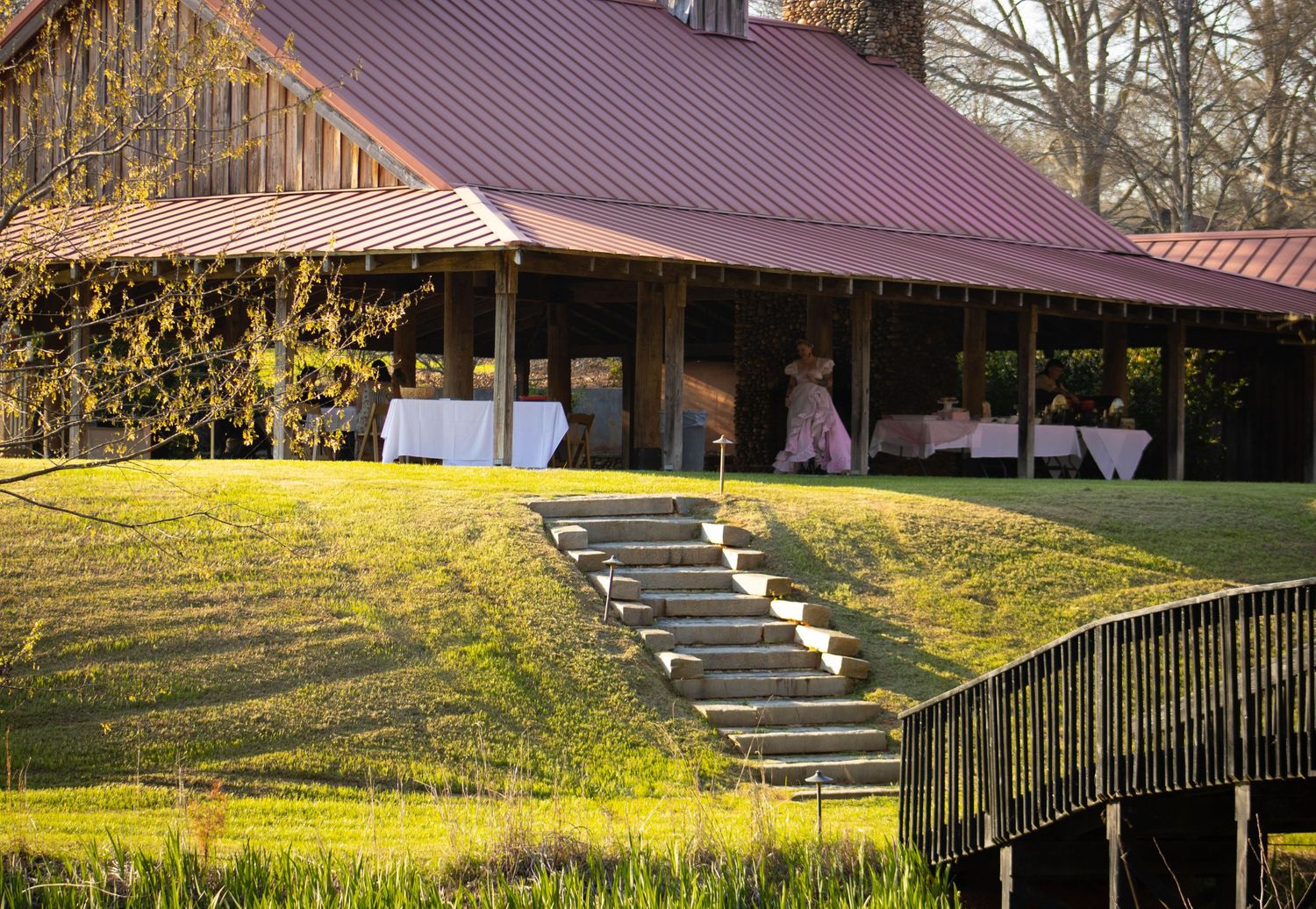 Rustic barn venue with red metal roof, stone steps, and elegant white-draped tables on a lush green hillside.