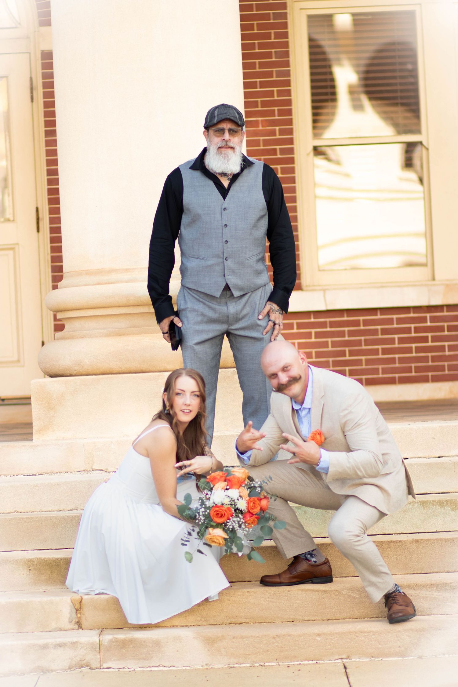 Bride and groom crouch playfully on steps with orange bouquet while officiant stands behind them.