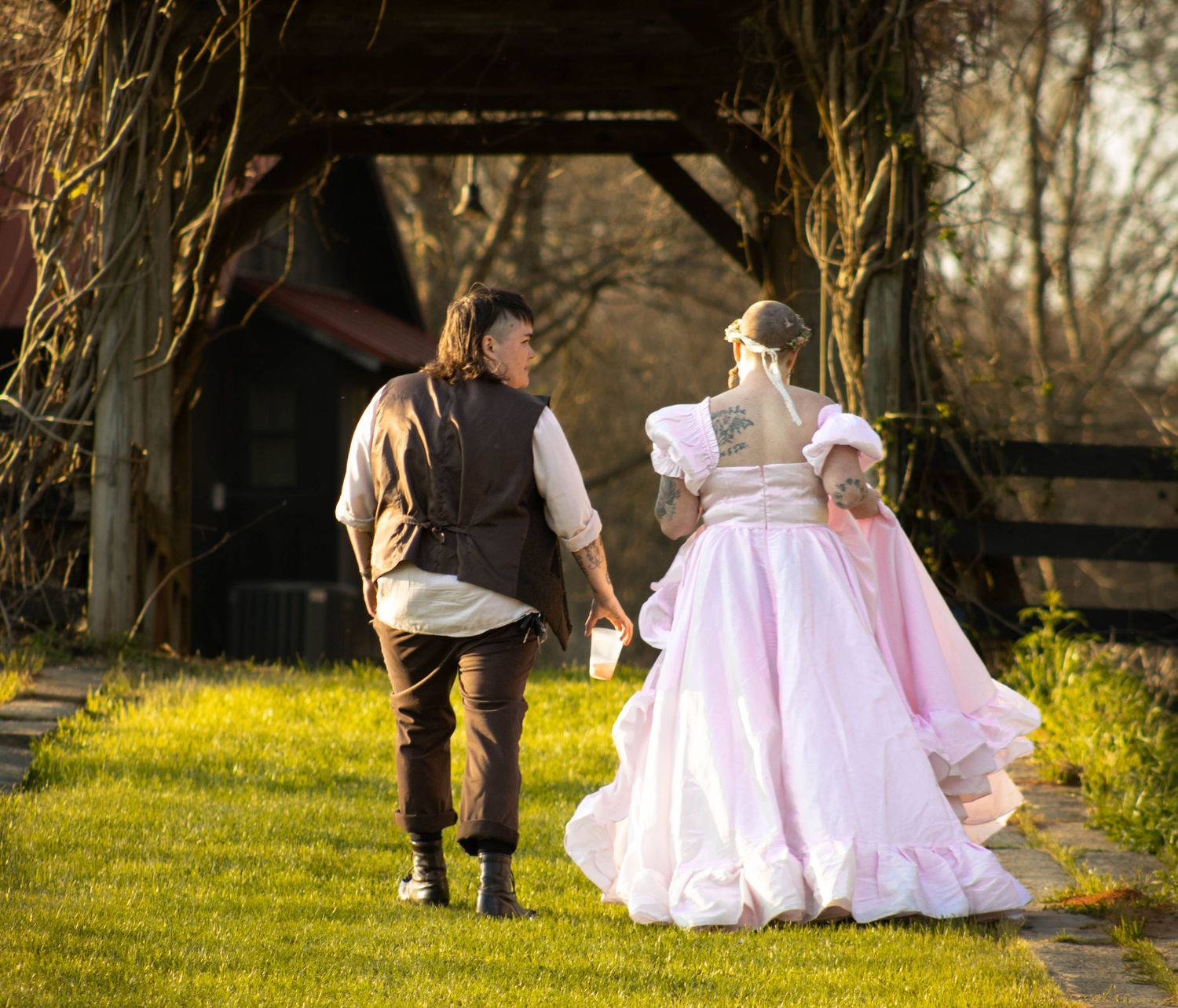 Couple in Renaissance costumes walking hand-in-hand through a sunlit garden path near a rustic wooden structure.