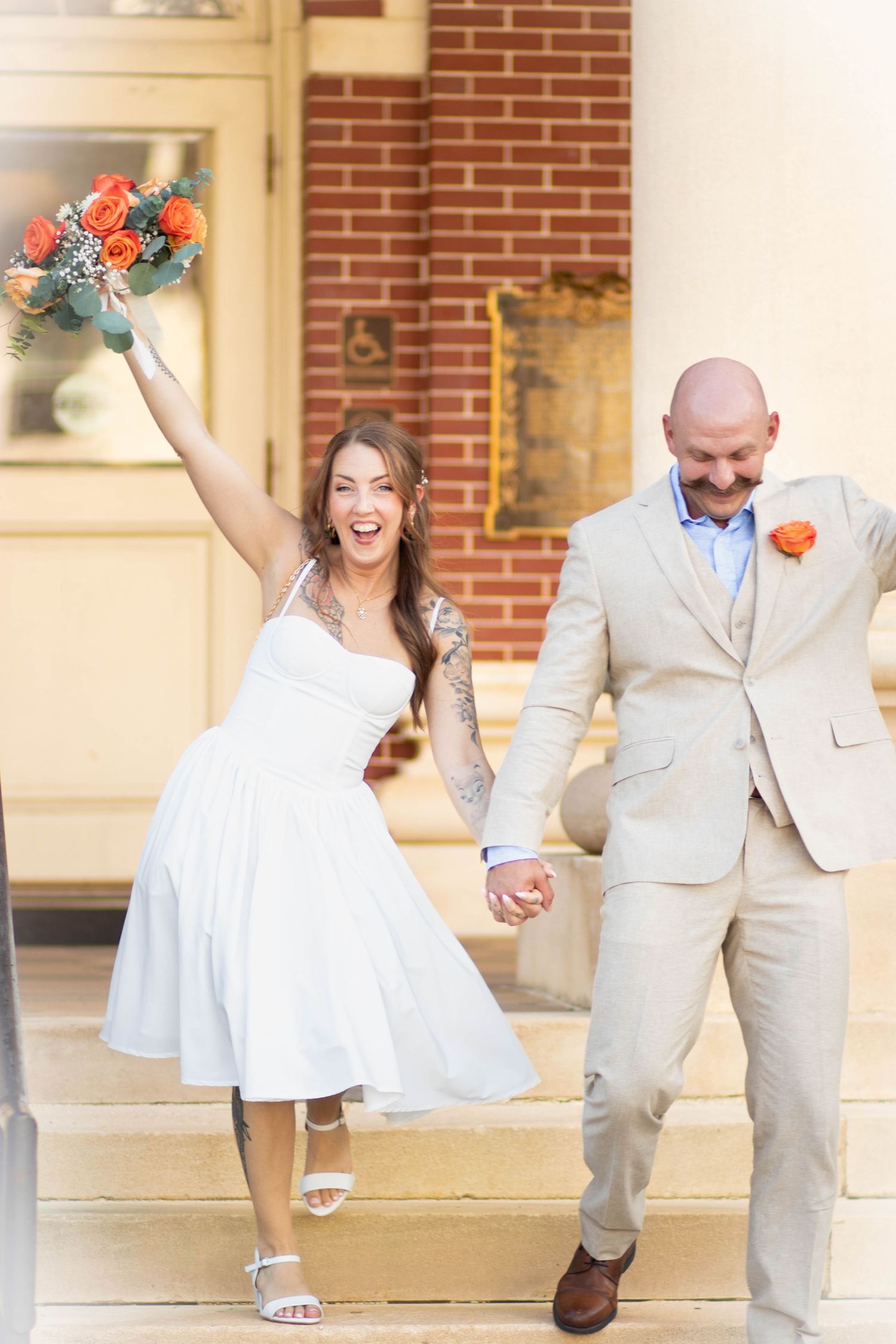 Joyful newlyweds exit venue steps, bride in white dress holding orange bouquet aloft, groom in tan suit with orange boutonniere.