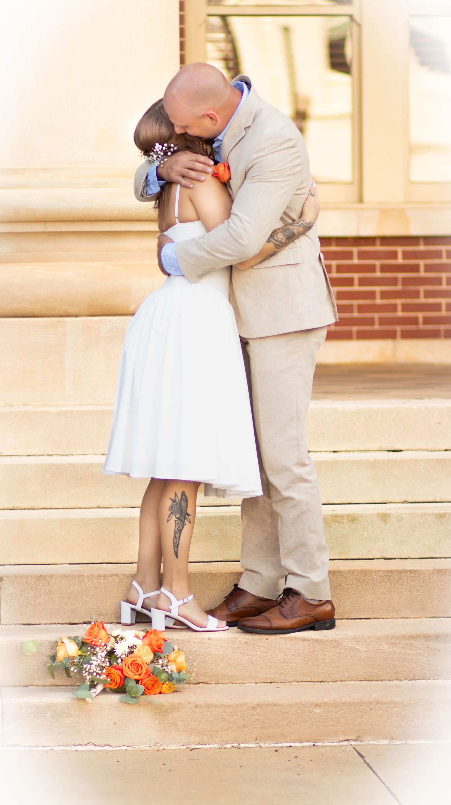 Newlywed couple sharing a kiss on stone steps, bride in white dress holding orange bouquet, groom in tan suit.