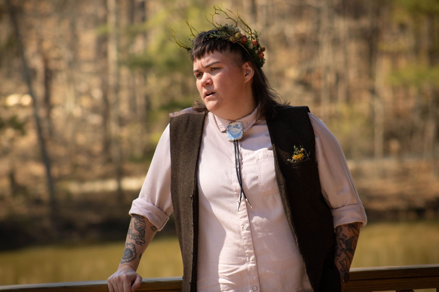 Person wearing a floral crown, bolo tie, and vest with tattoos, standing on a wooden bridge outdoors.