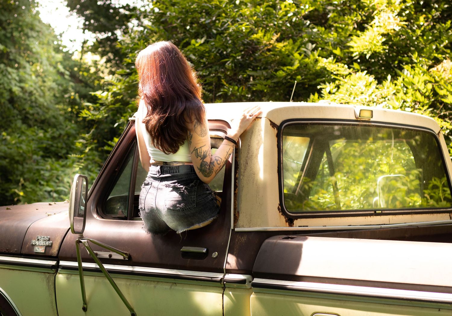 Woman with tattoos and red hair leaning out of a vintage truck door surrounded by lush green trees.