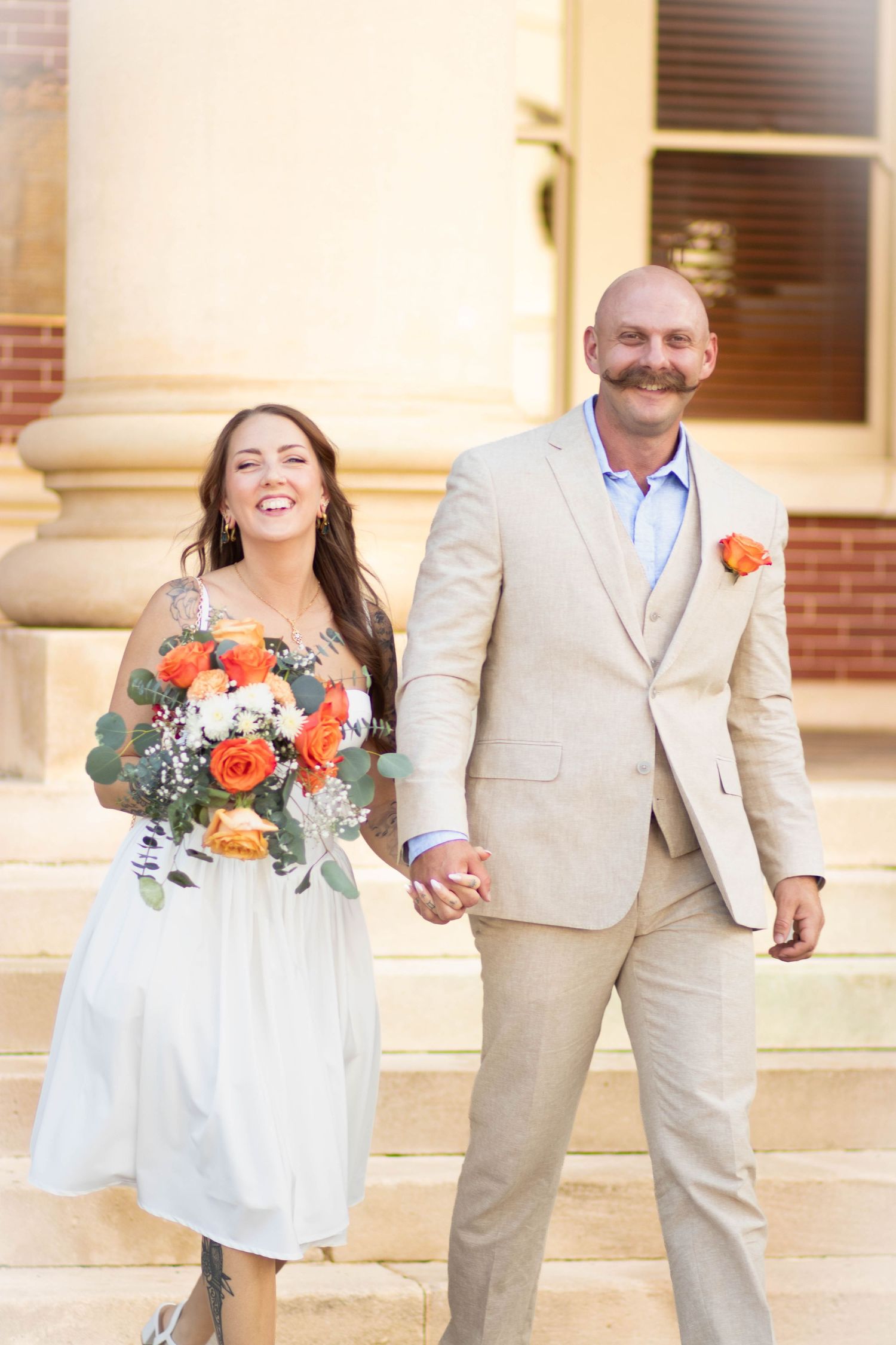 Smiling bride in white dress holding orange bouquet walks hand-in-hand with groom in tan suit on building steps.