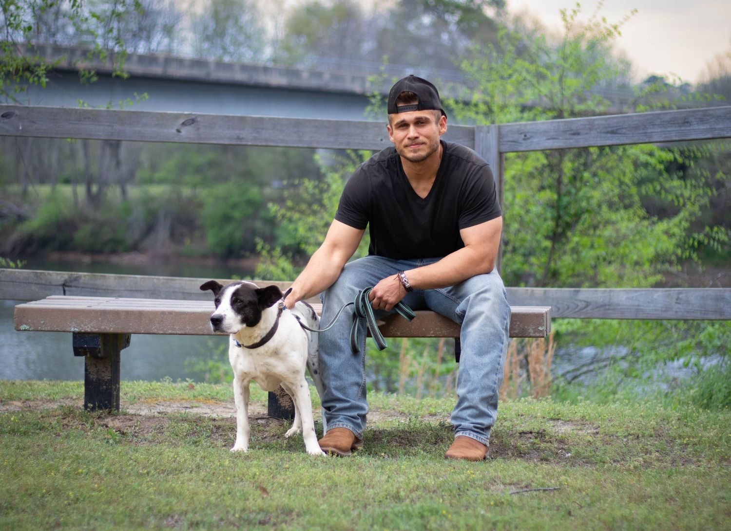 A man in a black shirt sits on a bench with his black and white dog in a park setting.