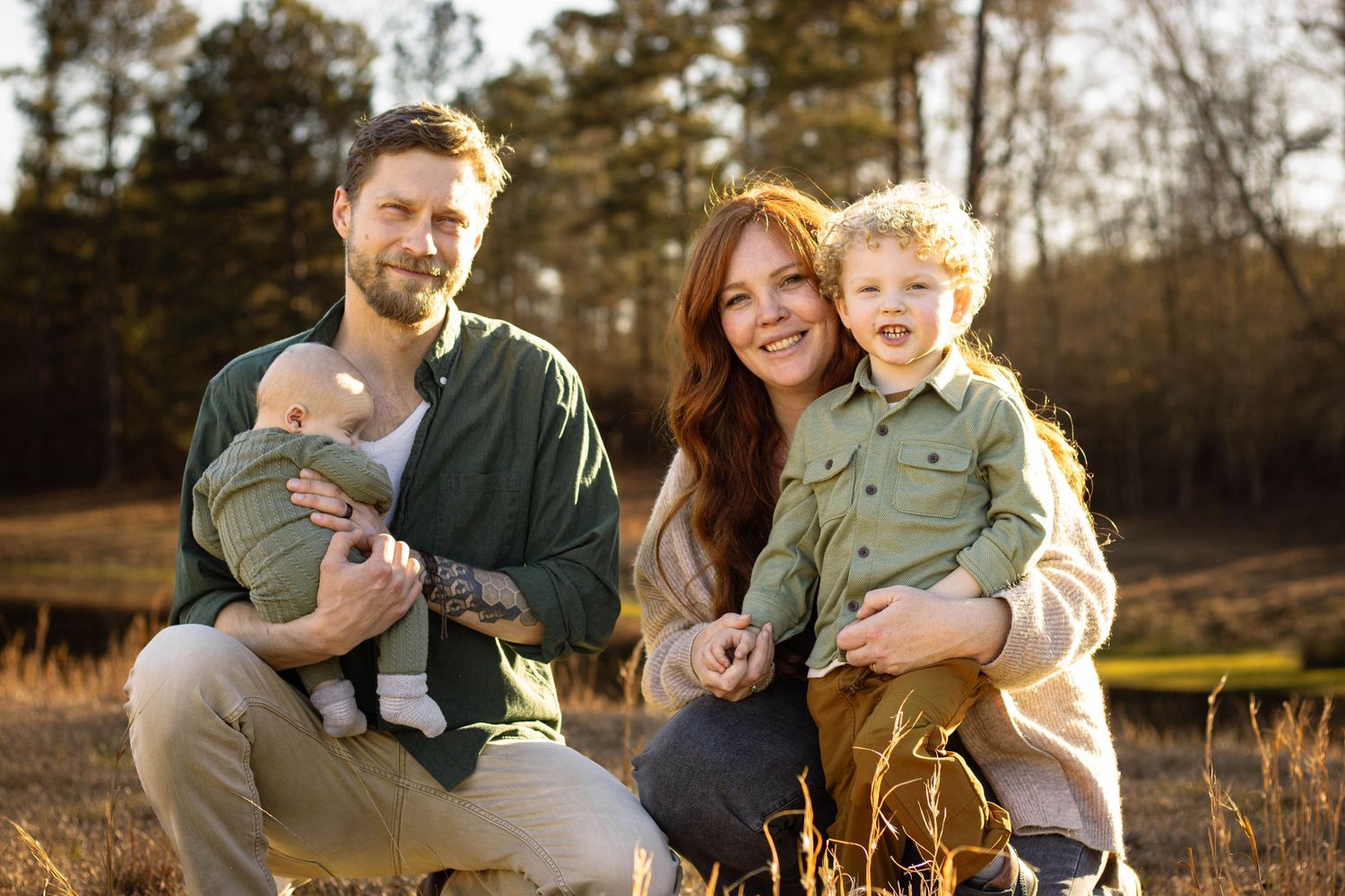 Happy family of four posing outdoors in golden hour light, parents holding two young children in a sunlit field.