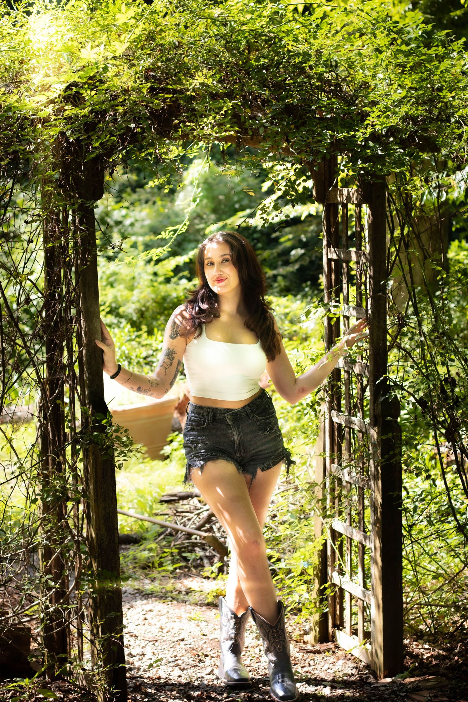 Young woman in white crop top, denim shorts, and cowboy boots posing under a rustic garden arbor surrounded by lush greenery.