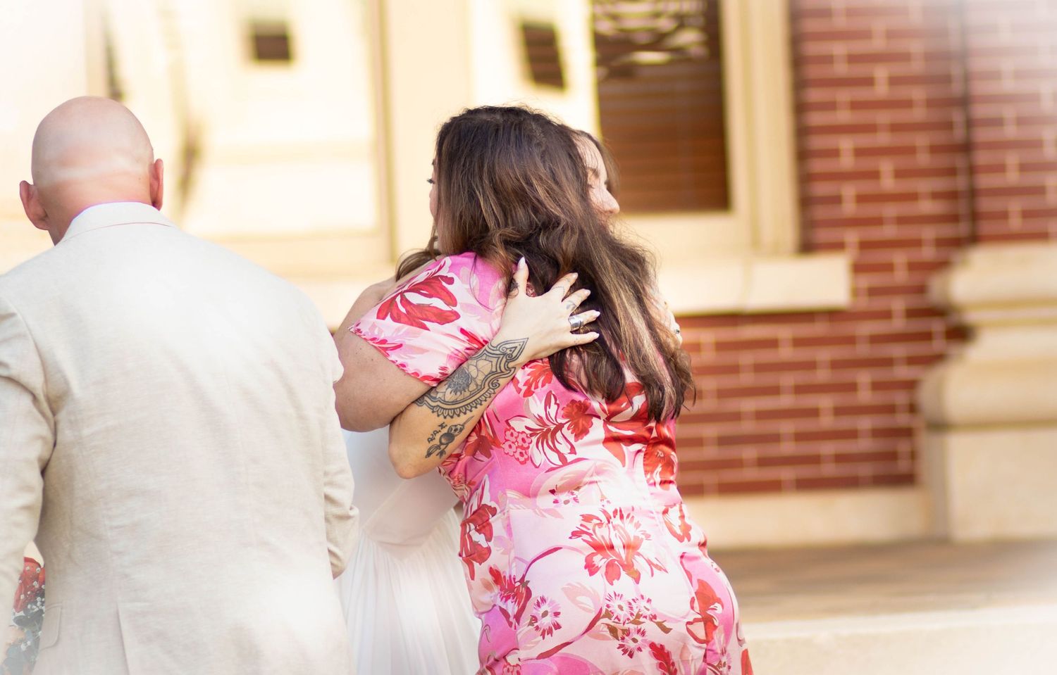 Two women in floral pink dresses embrace warmly outside a brick building, with a man in a white suit nearby.