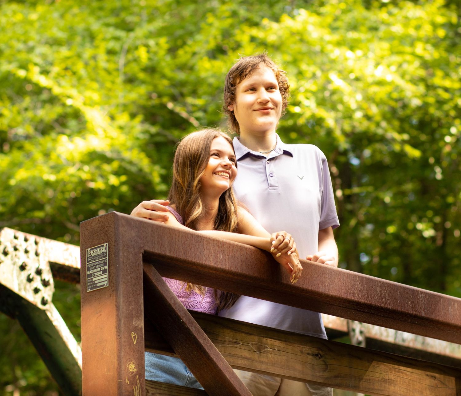 A young couple smiles while leaning on a wooden railing surrounded by lush green trees outdoors.