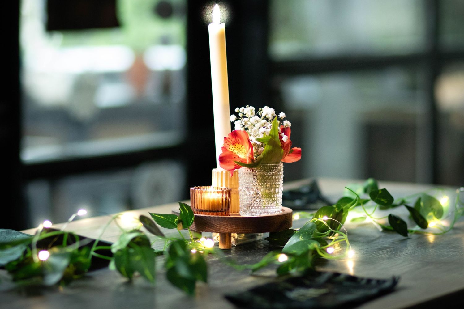 Elegant wedding centerpiece with tall candle, small flowers, fairy lights, and green ivy on a wooden table.