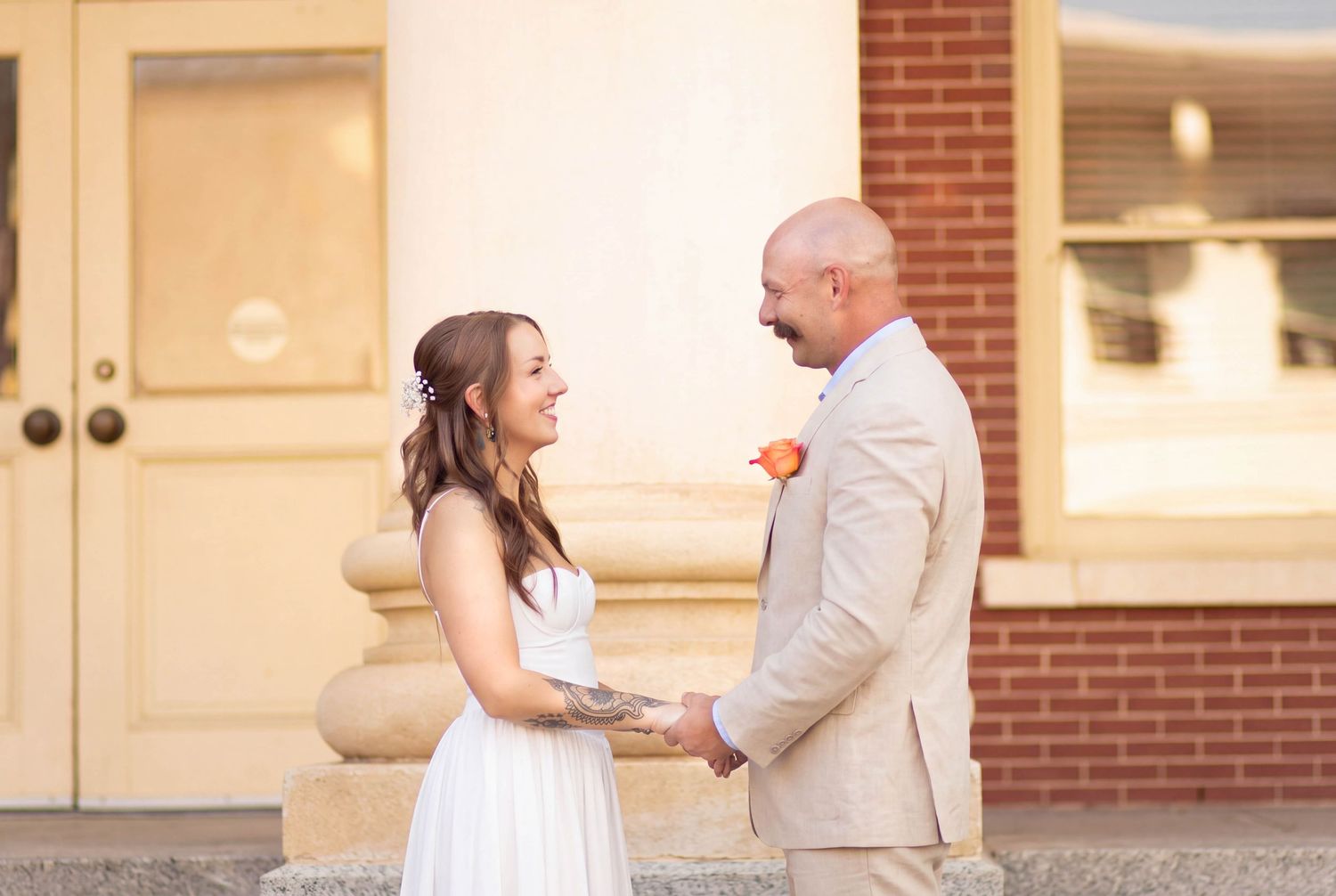 A bride and groom hold hands and smile at each other outdoors near a brick building on their wedding day.