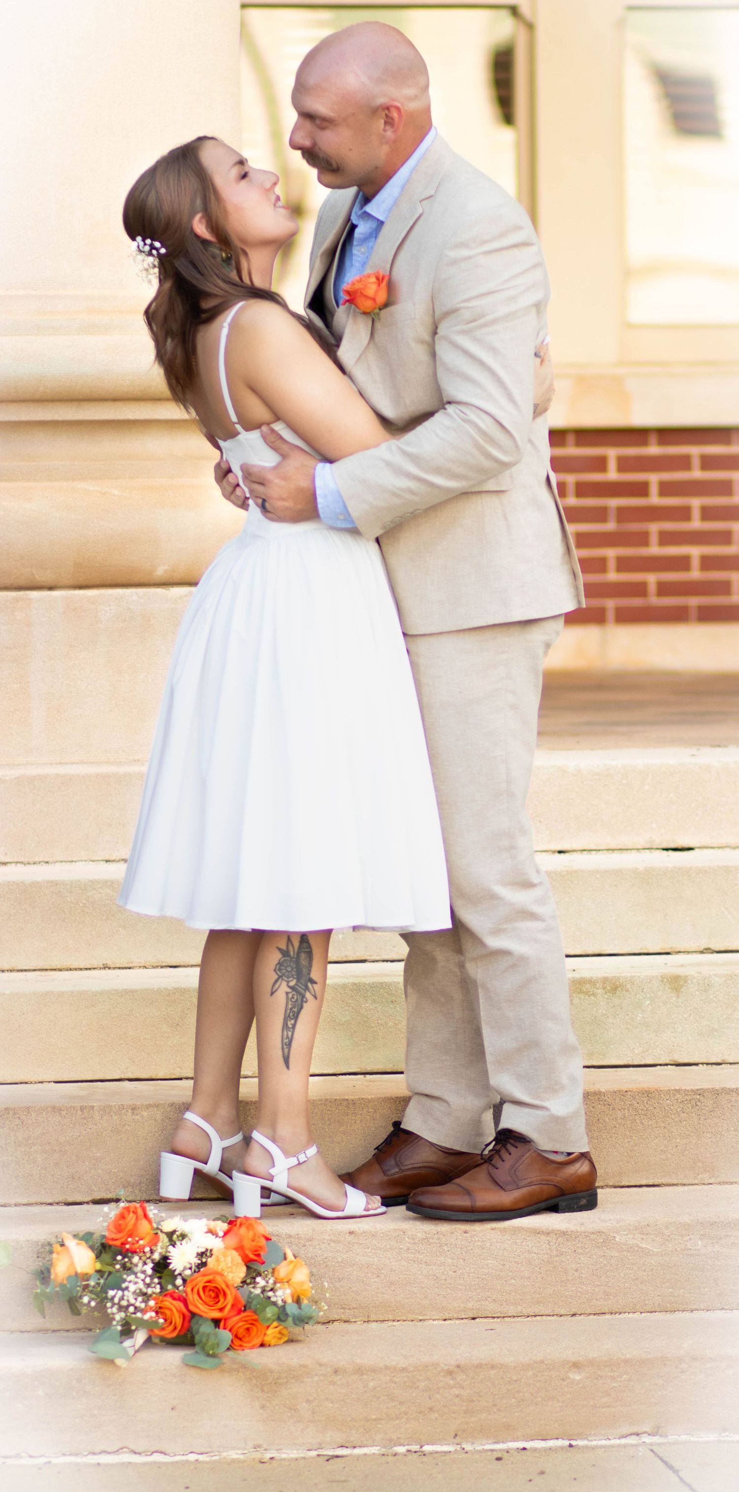 Bride and groom embrace on stone steps, she in white dress with bouquet, he in tan suit with orange boutonniere.