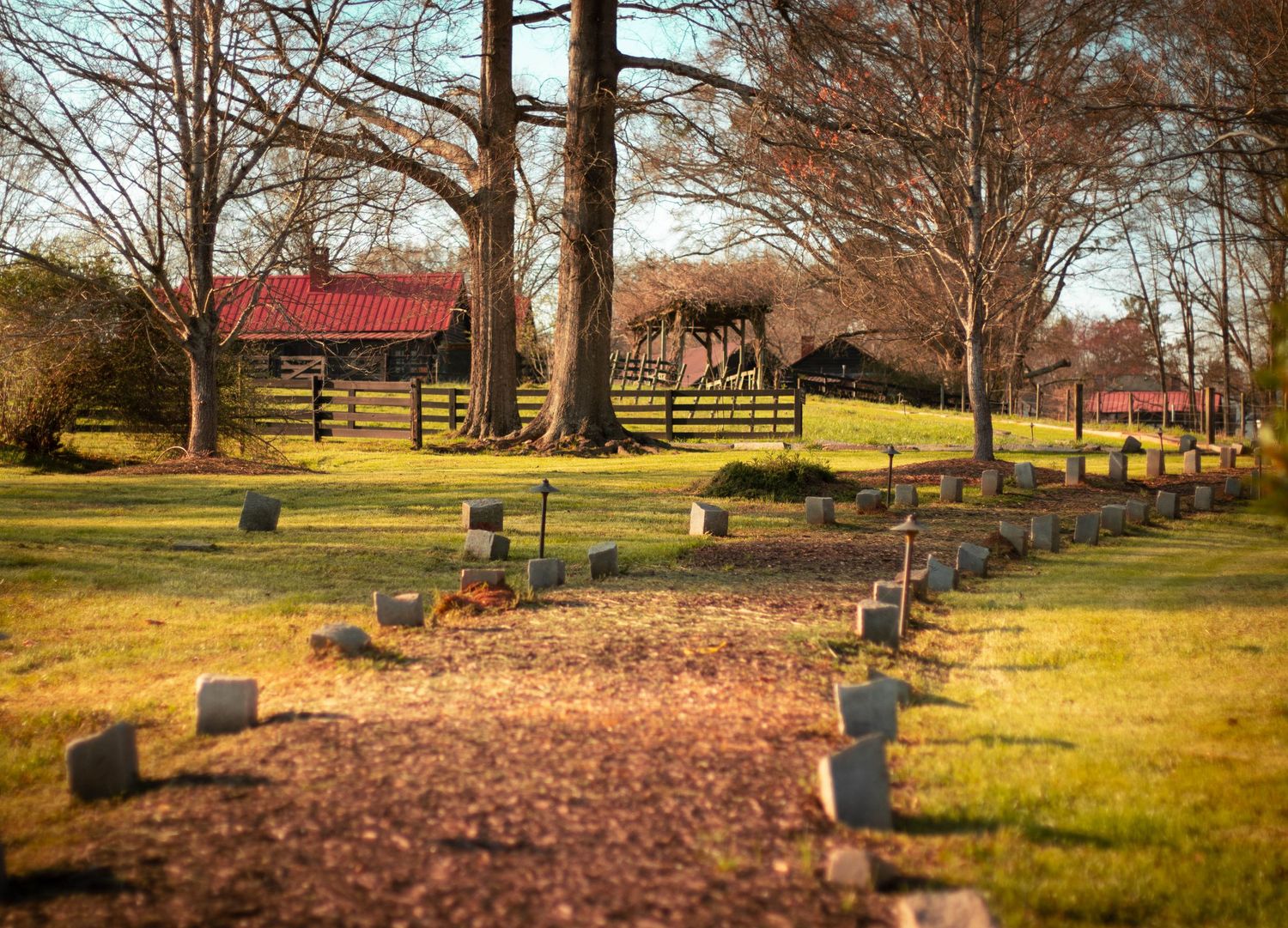 Historic cemetery with rows of grave markers on green grass, surrounded by bare trees and a red-roofed barn in warm afternoon light.
