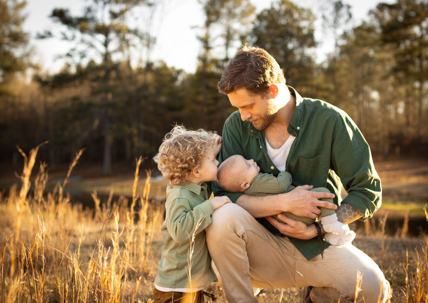 A father kneels in a golden field, tenderly holding a newborn while his young curly-haired child looks on.