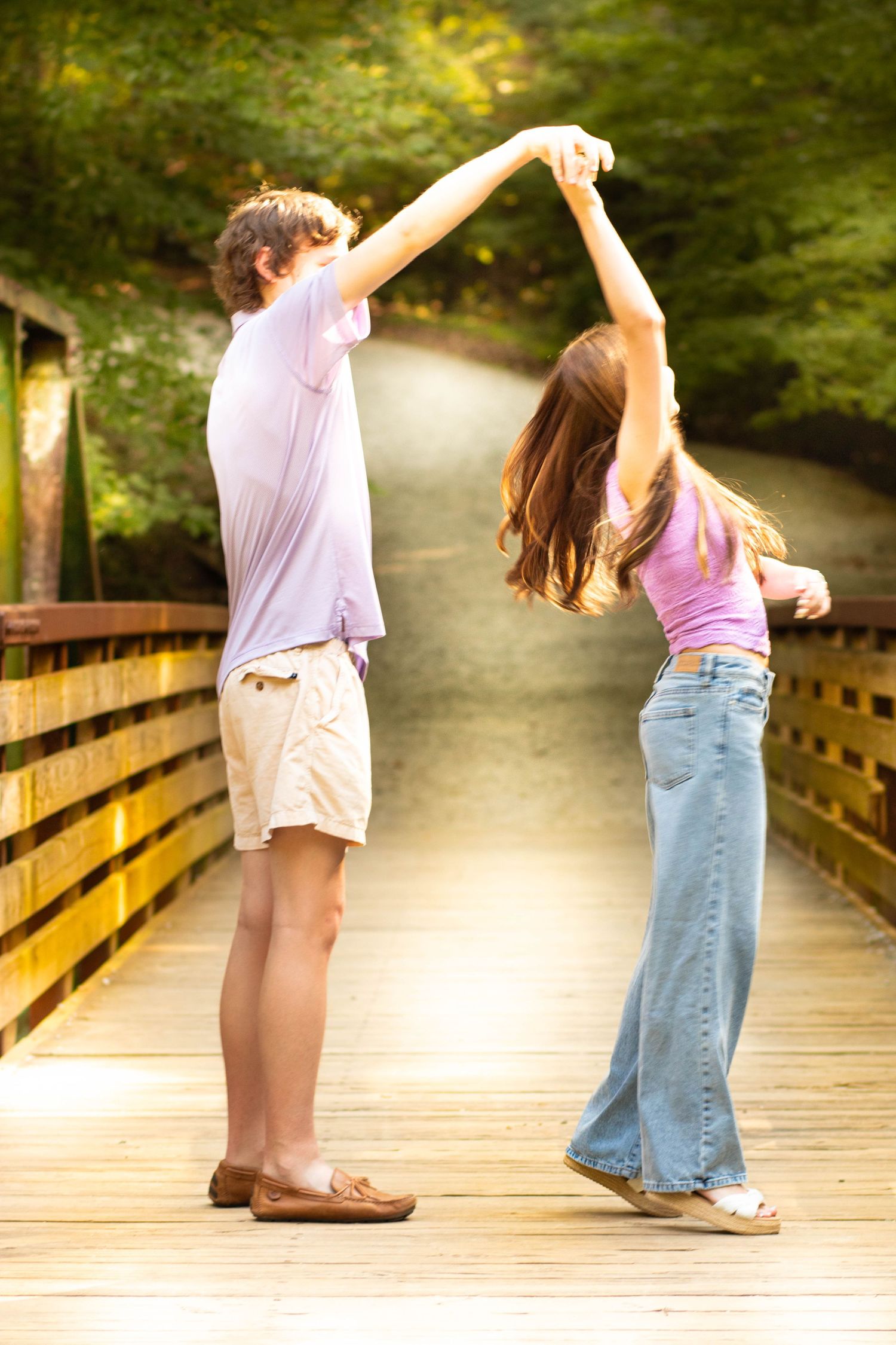 A young couple dancing together on a wooden bridge surrounded by lush green trees in a park.