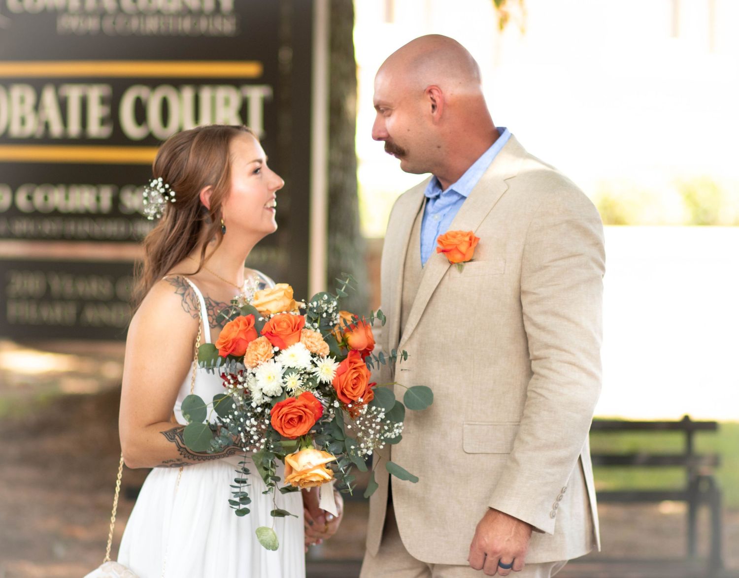 Bride and groom smiling at each other outside probate court, bride holding orange rose bouquet, groom in tan suit.
