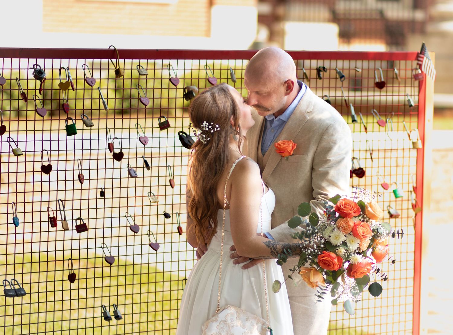 Bride and groom sharing a kiss in front of a decorative fence adorned with colorful locks at their wedding.
