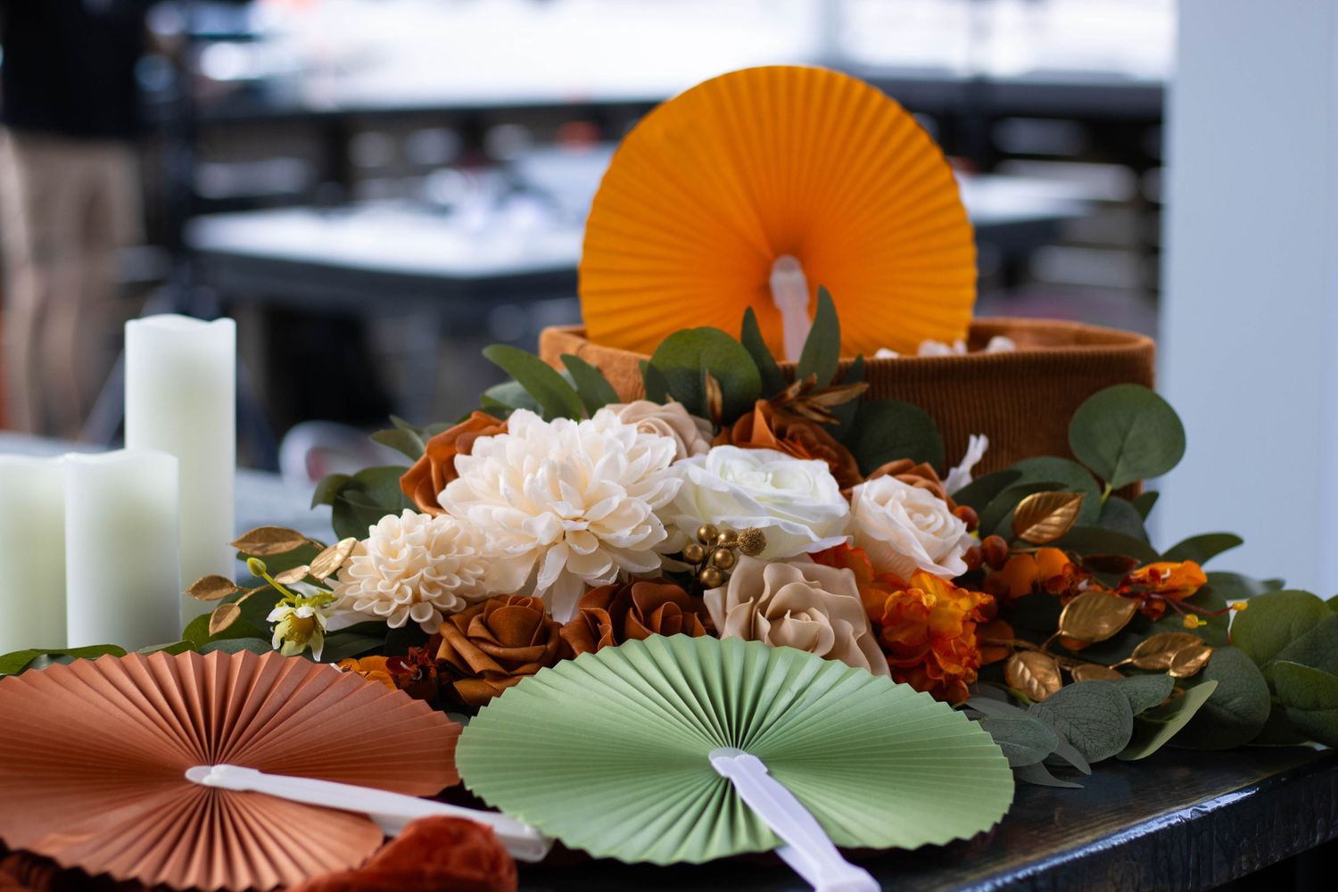 Elegant wedding table centerpiece with white and orange flowers, paper fans, and candles in autumn colors.