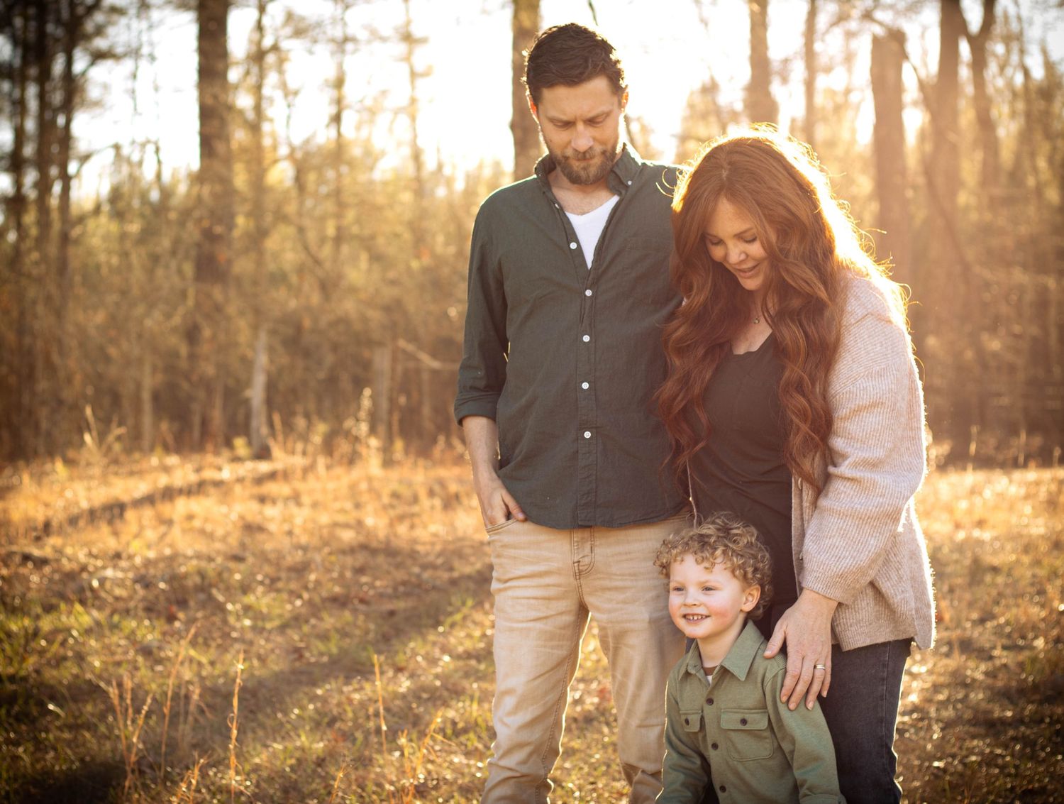 Family of three sharing tender moment outdoors, parents looking down at young boy in warm golden forest light.