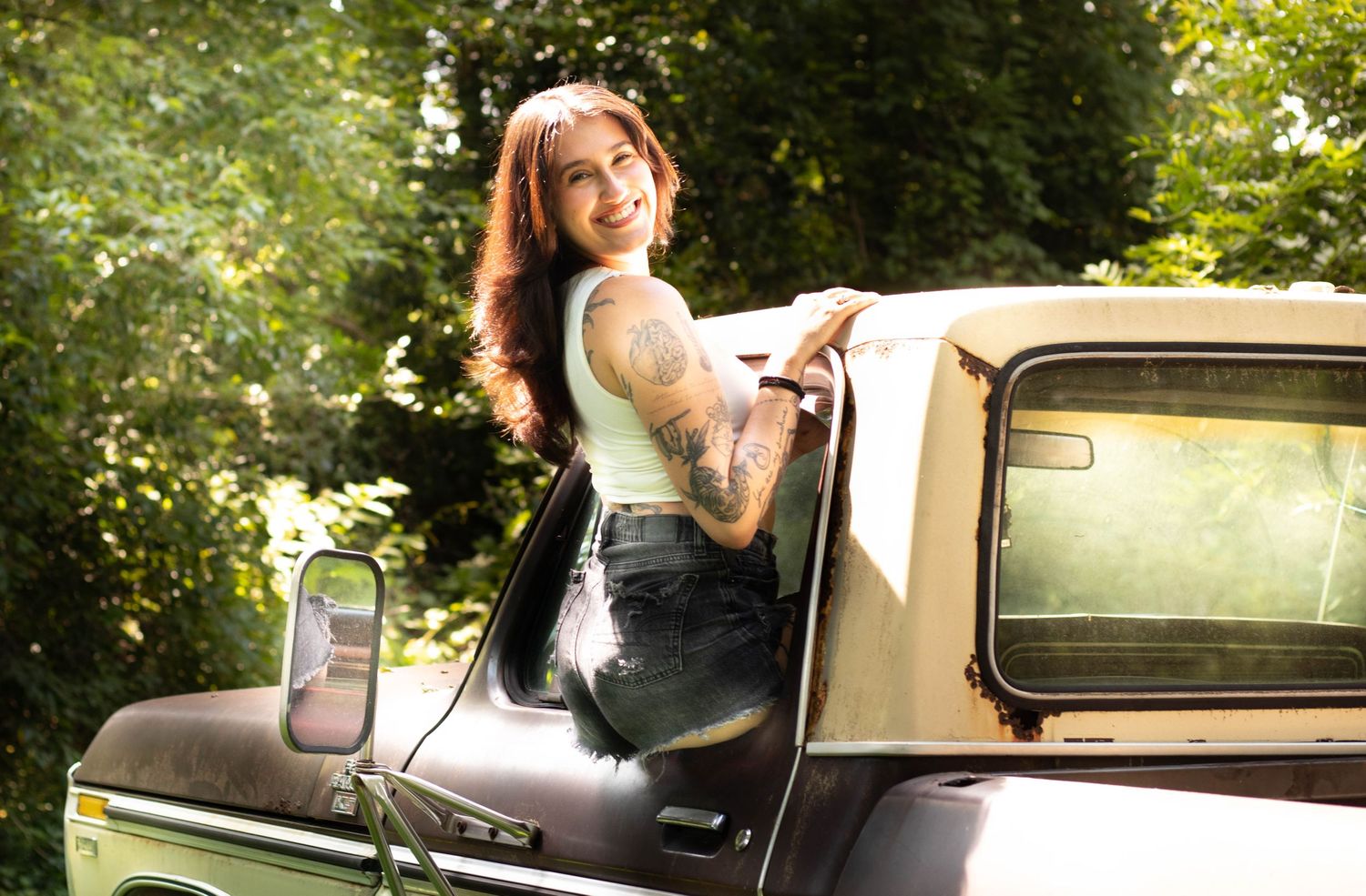Smiling woman with tattoos and red hair posing beside a vintage tan truck outdoors surrounded by lush green trees.