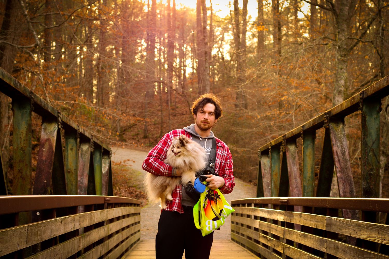 Man in plaid shirt holding a fluffy dog on a wooden bridge surrounded by autumn forest trees.