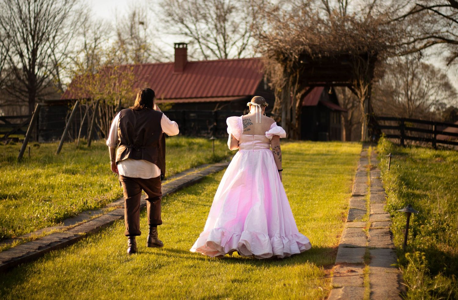 A bride in a pink ball gown and groom walk hand in hand toward a rustic farm building.