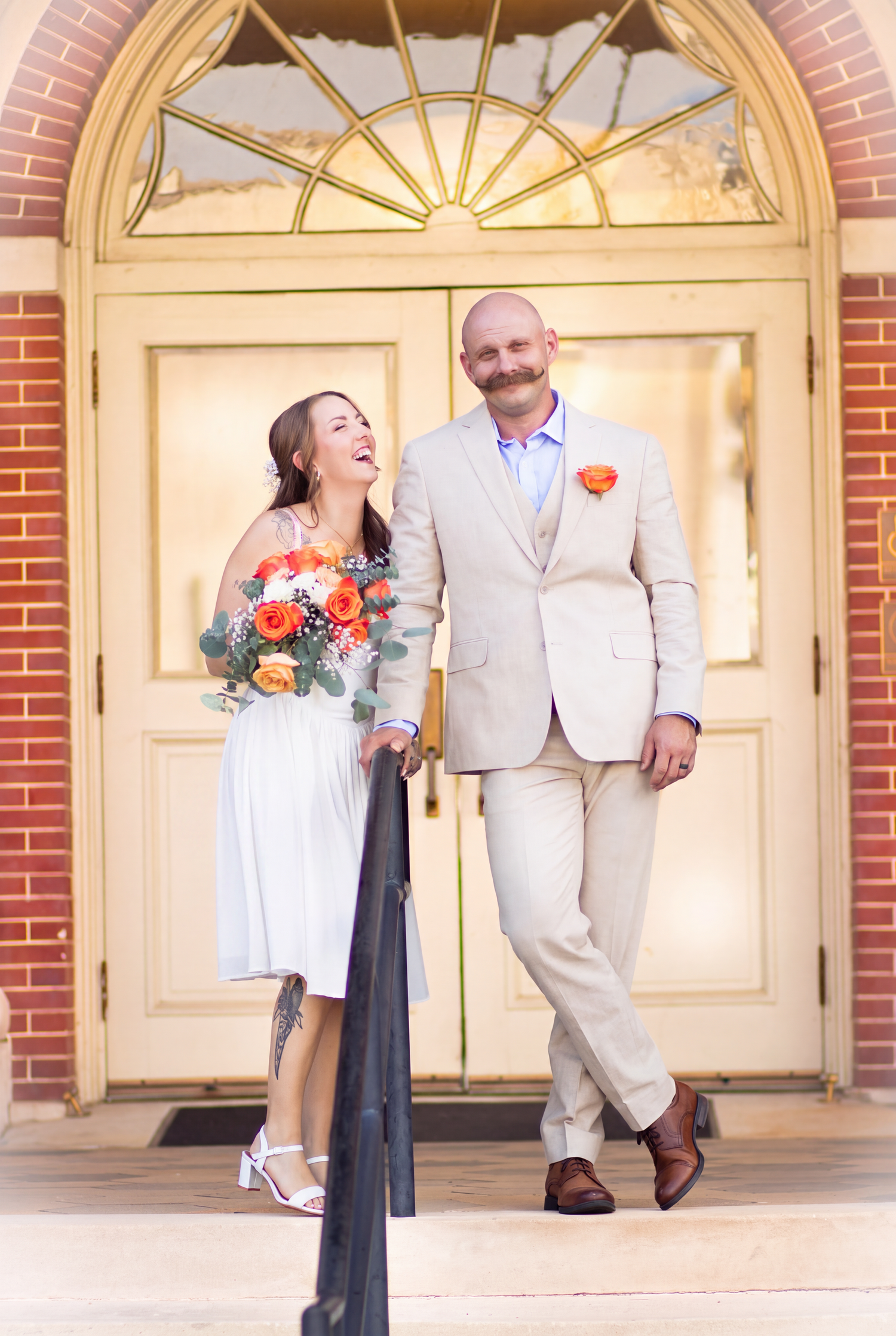 Happy couple in wedding attire laughing on church steps, bride holding orange bouquet, groom in tan suit with orange boutonniere.