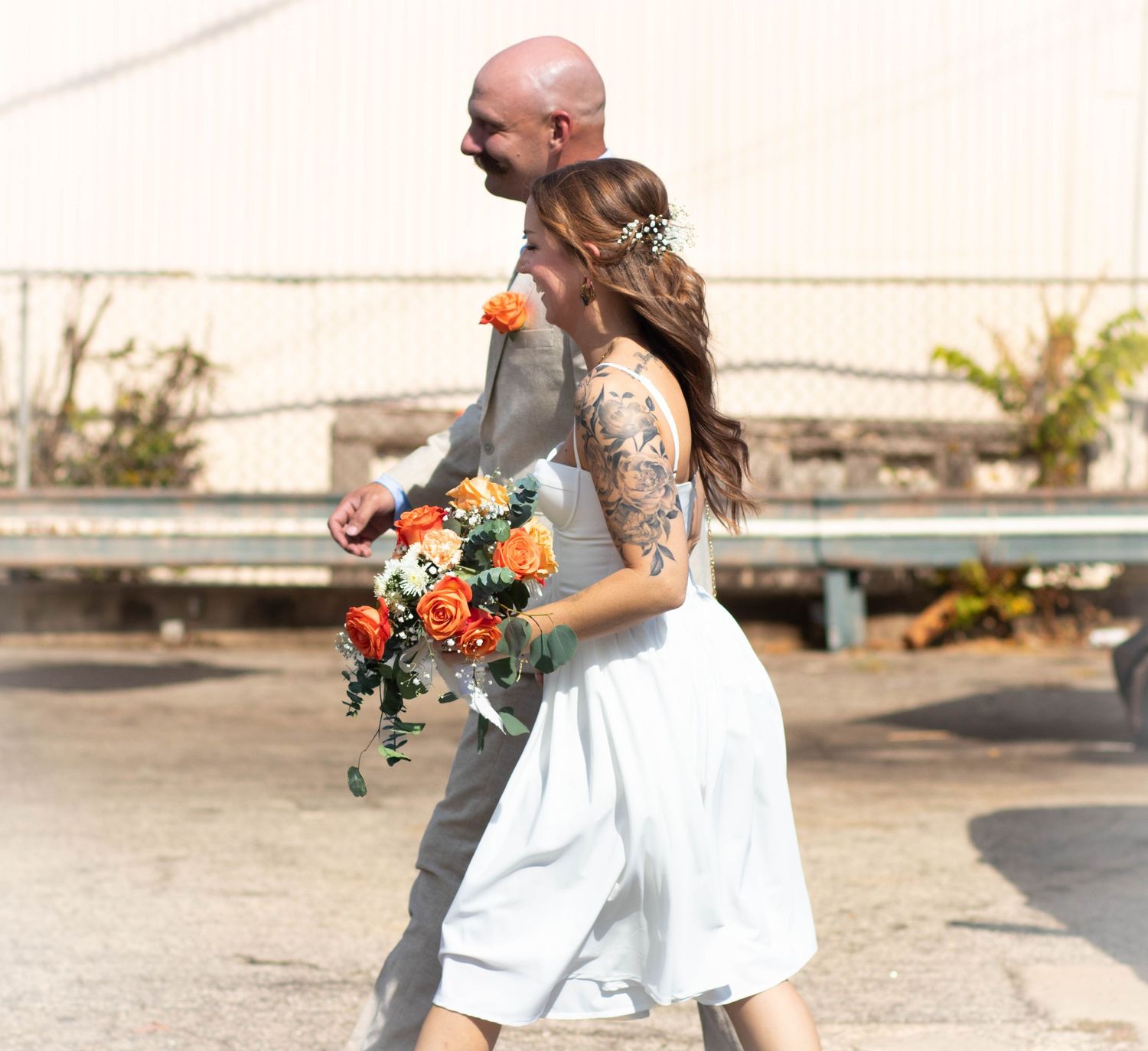 Bride in white dress holding orange rose bouquet walks with tattooed groom outdoors on their wedding day.