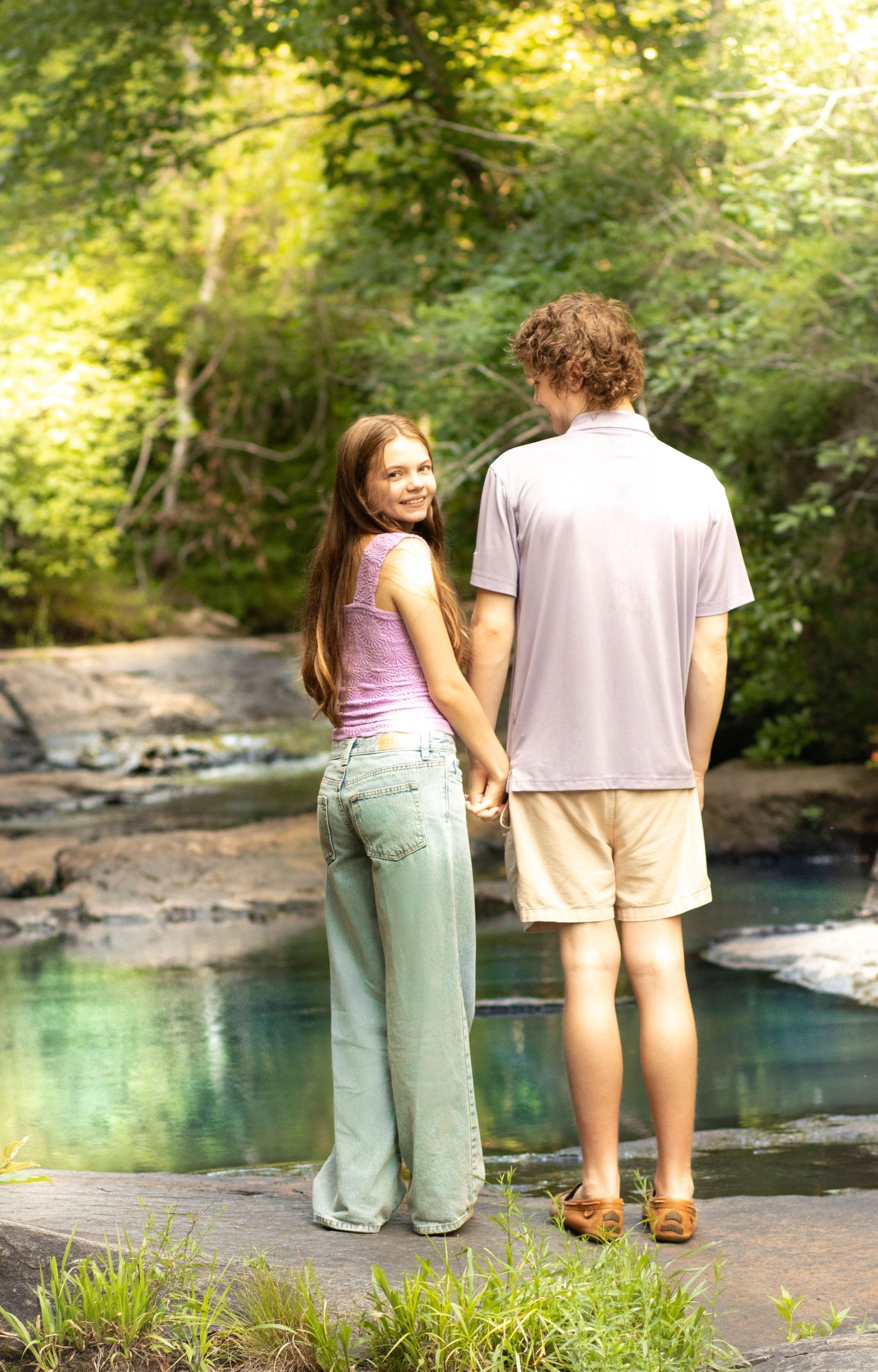 Young couple holding hands by a serene forest creek, girl smiling back at camera in casual summer attire.