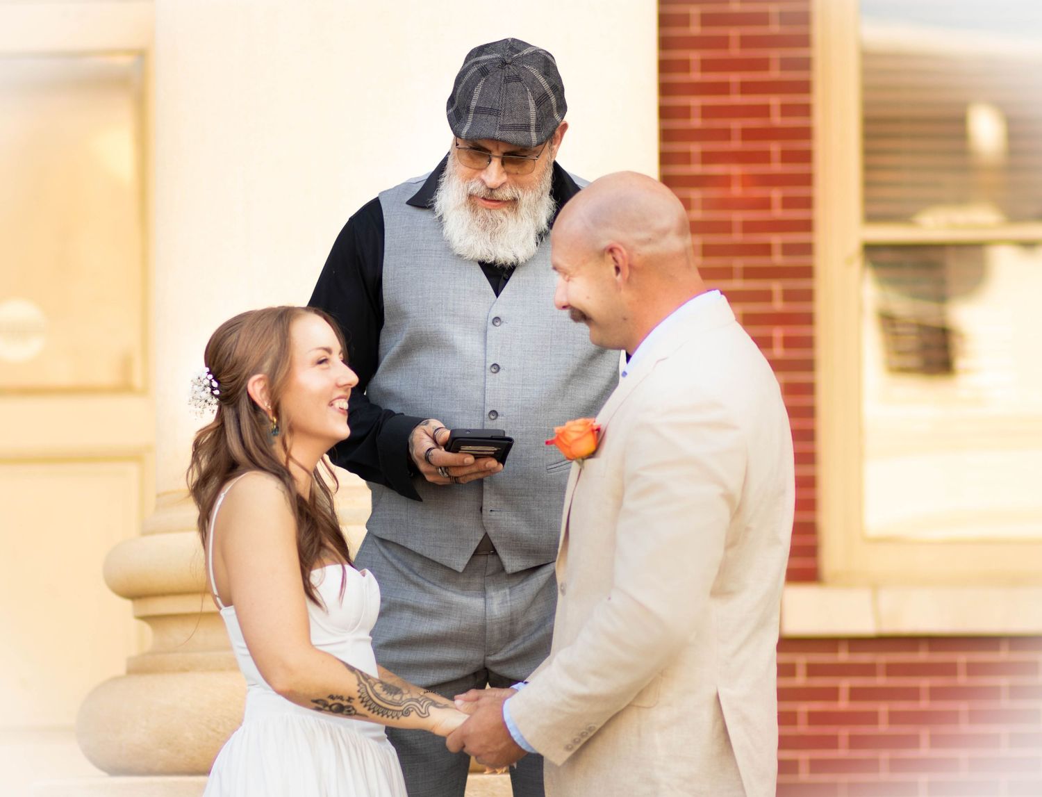 Bride and groom holding hands during outdoor wedding ceremony with bearded officiant reading from phone.