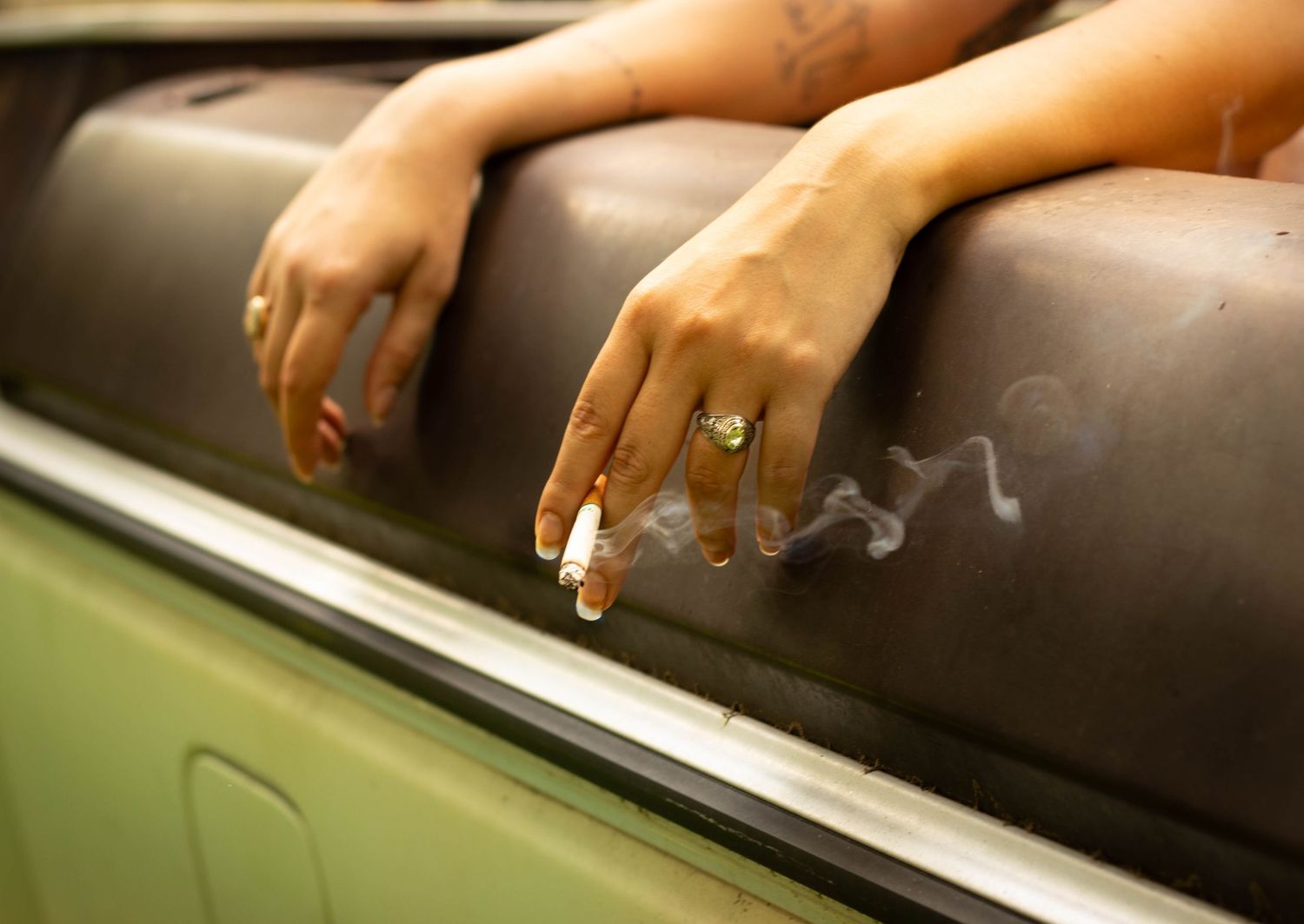 Close-up of a woman's hands resting on a leather seat, holding a lit cigarette with smoke rising, wearing a green ring.