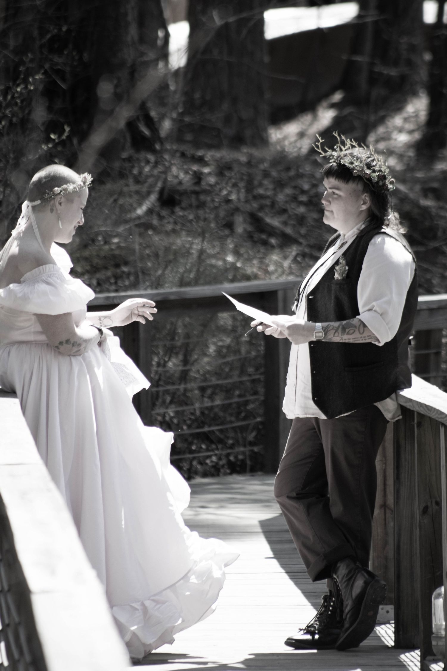 Black and white photo of two brides exchanging vows on a wooden bridge, one in a gown, one in a vest with floral crown.