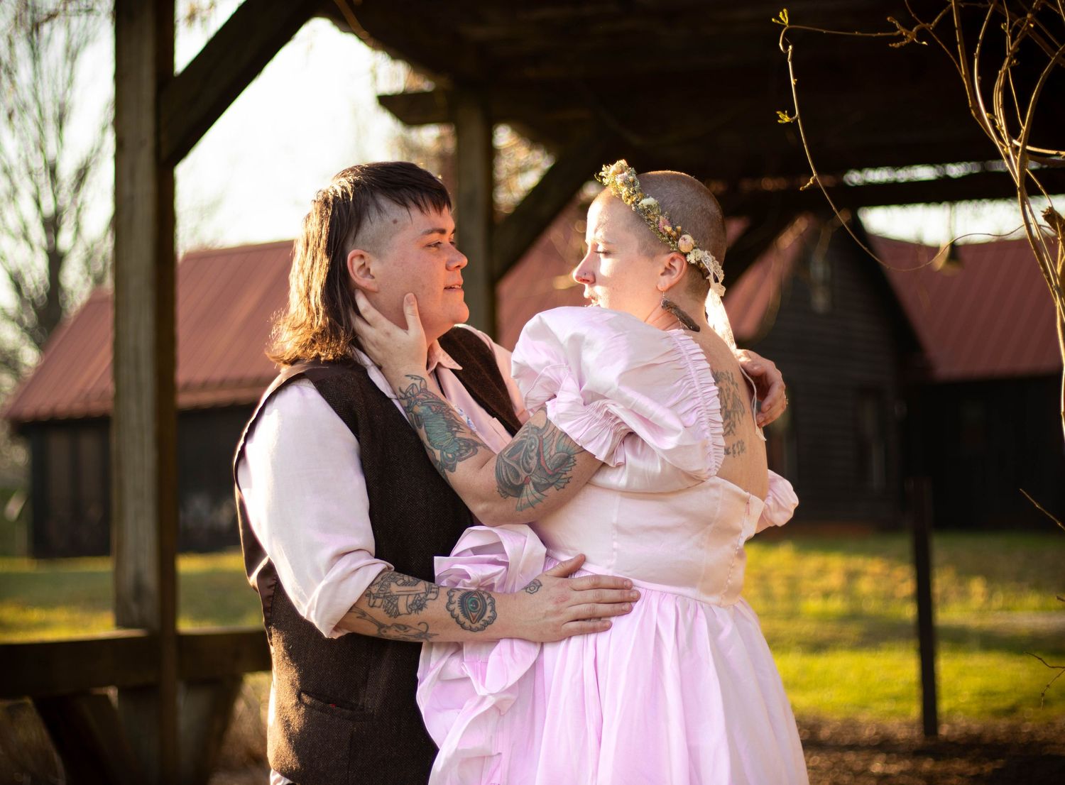 Couple in wedding attire embracing outdoors, bride in pink gown, groom with tattoos in vest, rustic barn background.