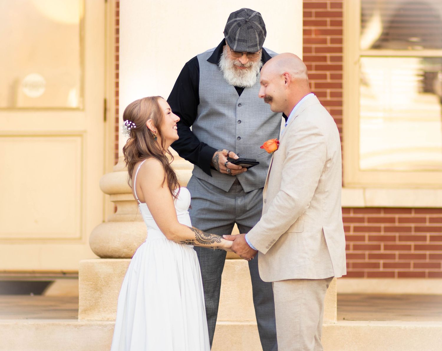 Bride and groom hold hands during outdoor wedding ceremony as bearded officiant reads from phone.