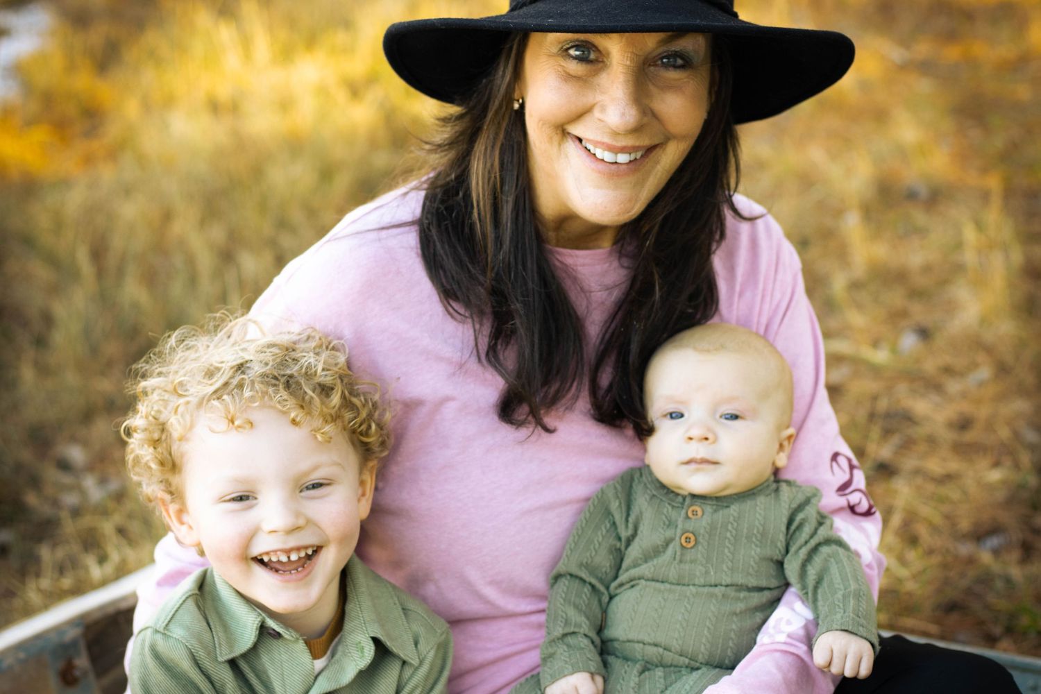 Smiling mother in black hat with two young children outdoors in autumn setting.