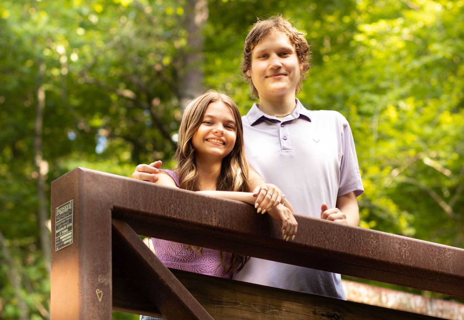 Two smiling teens pose on a wooden bridge surrounded by lush green trees in a scenic outdoor setting.