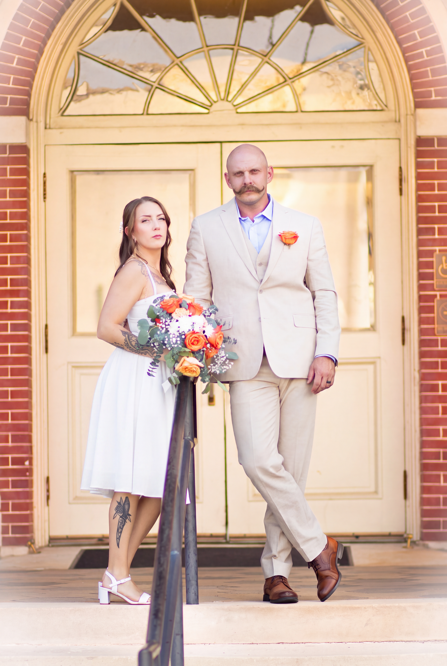 Bride in white dress holding orange bouquet stands with groom in tan suit before elegant yellow doors at wedding venue.