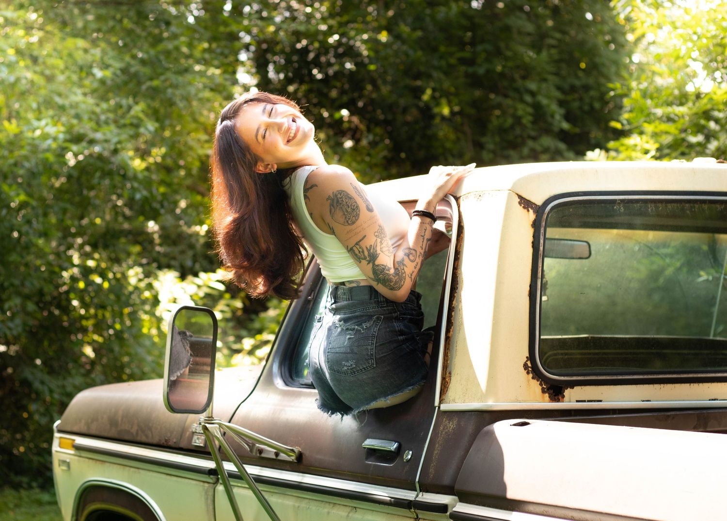 Tattooed woman leaning out of vintage truck window, smiling joyfully, surrounded by lush green trees.