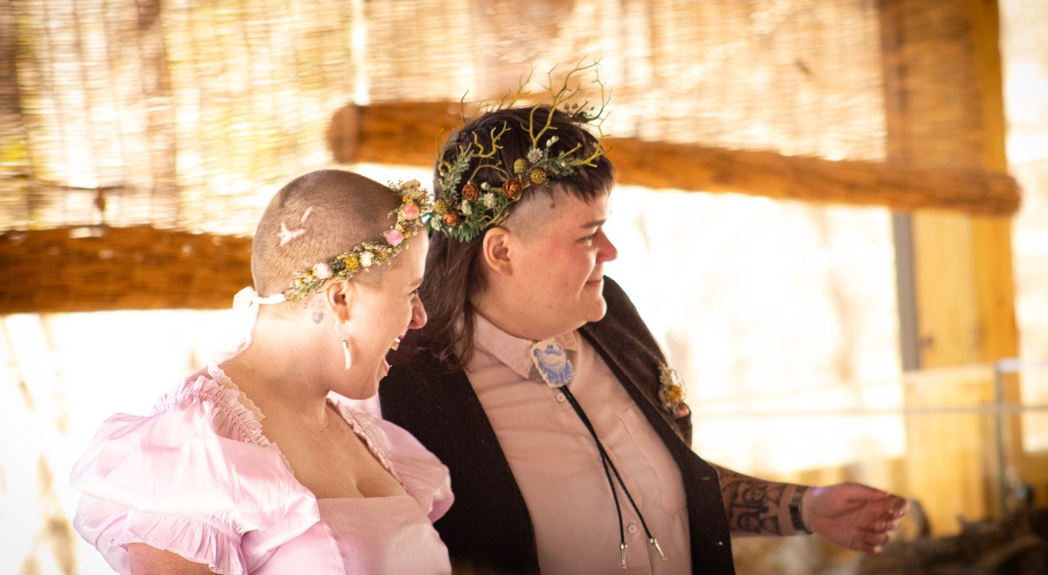 Two people wearing floral crowns share a tender moment at an outdoor wedding ceremony.