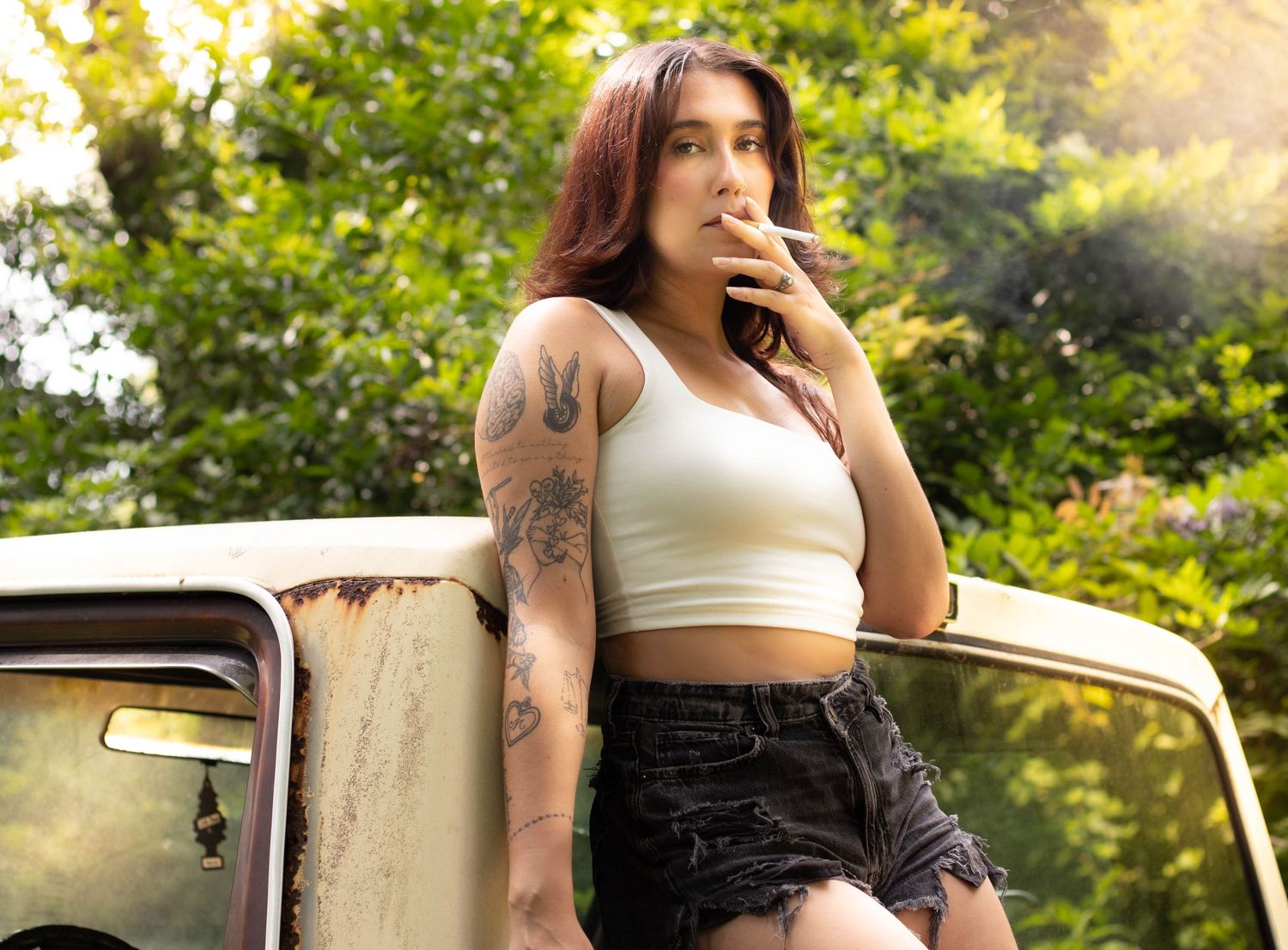 Tattooed woman in white crop top and denim shorts leaning on vintage truck outdoors.