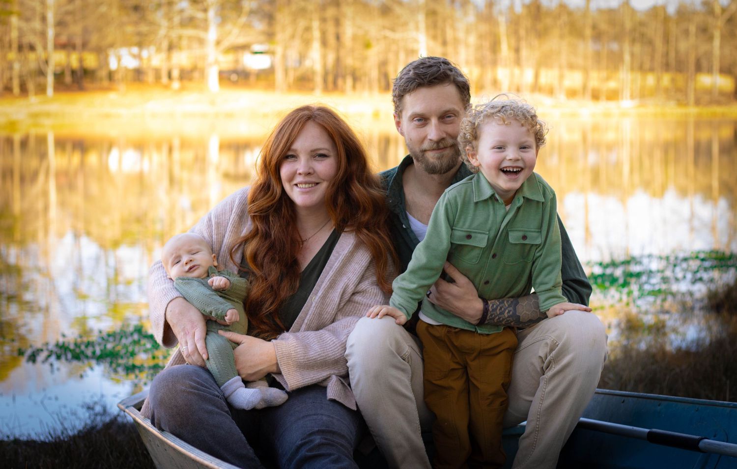 Happy family of three sitting together near sunlit pond with golden reflections in background.