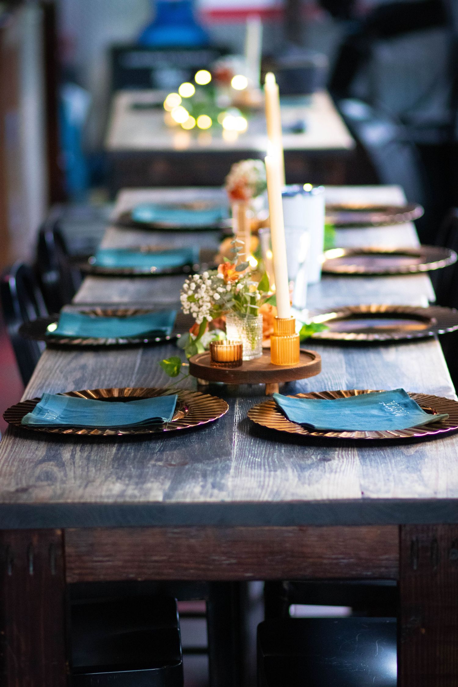 Elegant rustic wooden dining table set with teal napkins, gold charger plates, candles, and floral centerpieces.