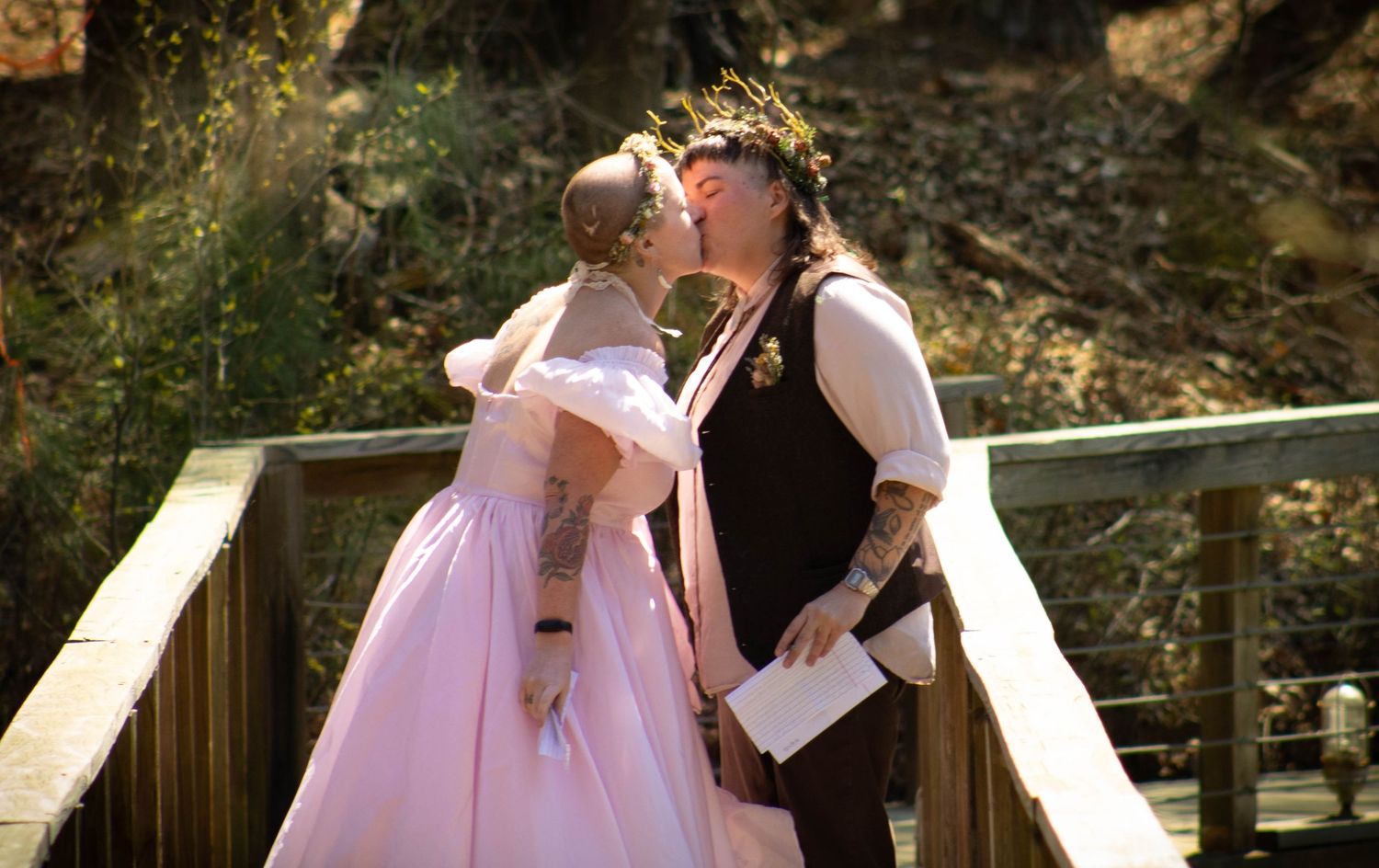 Couple sharing a kiss at outdoor wedding, bride in pink gown, groom in medieval attire on wooden bridge.