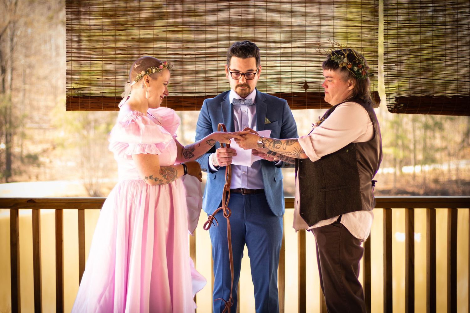 Couple exchanges rings during outdoor wedding ceremony with officiant in pink dress and brown suit.